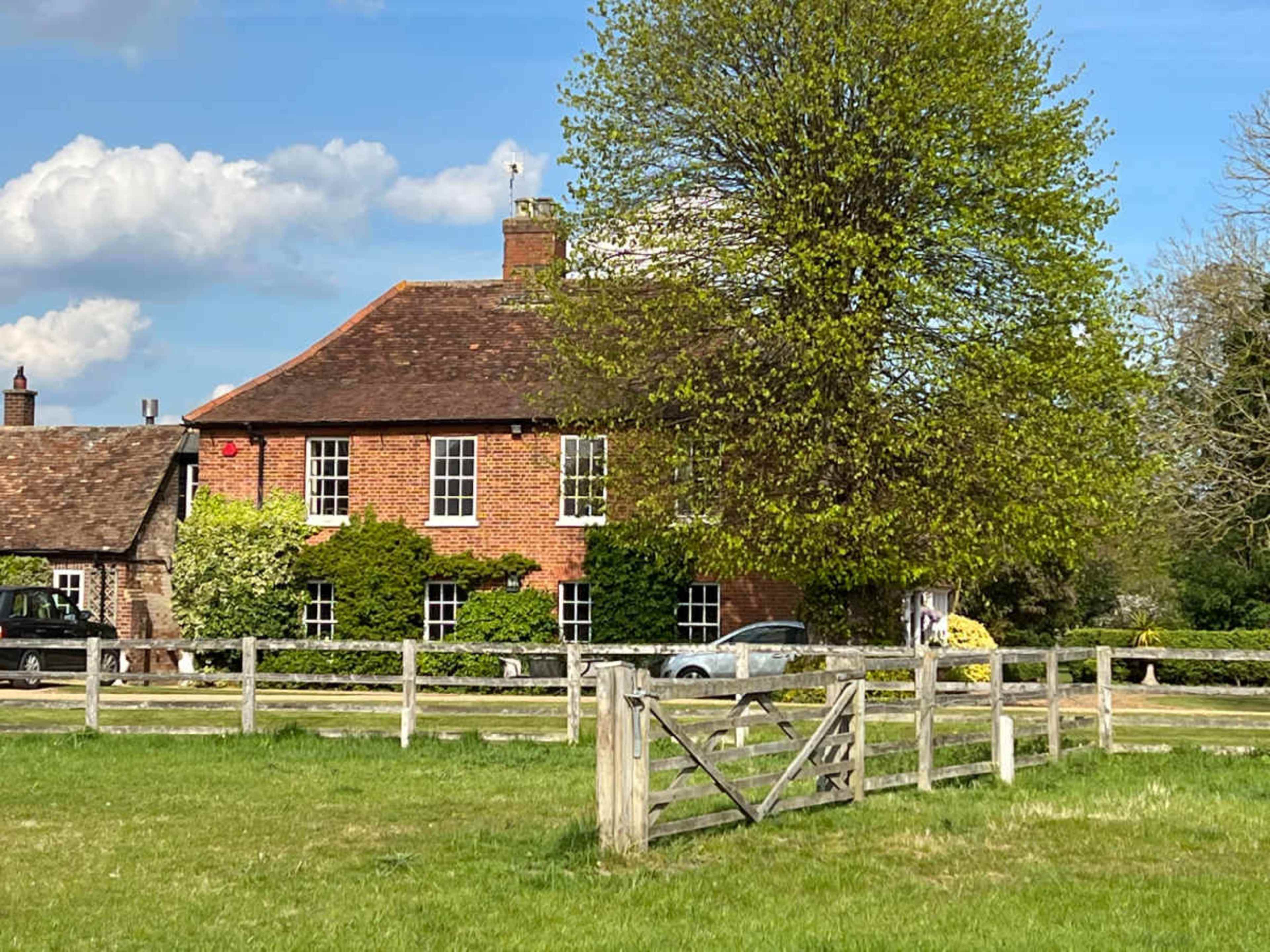 A brick house with a sloped roof is surrounded by green trees and a grassy field, with a wooden fence in the foreground.