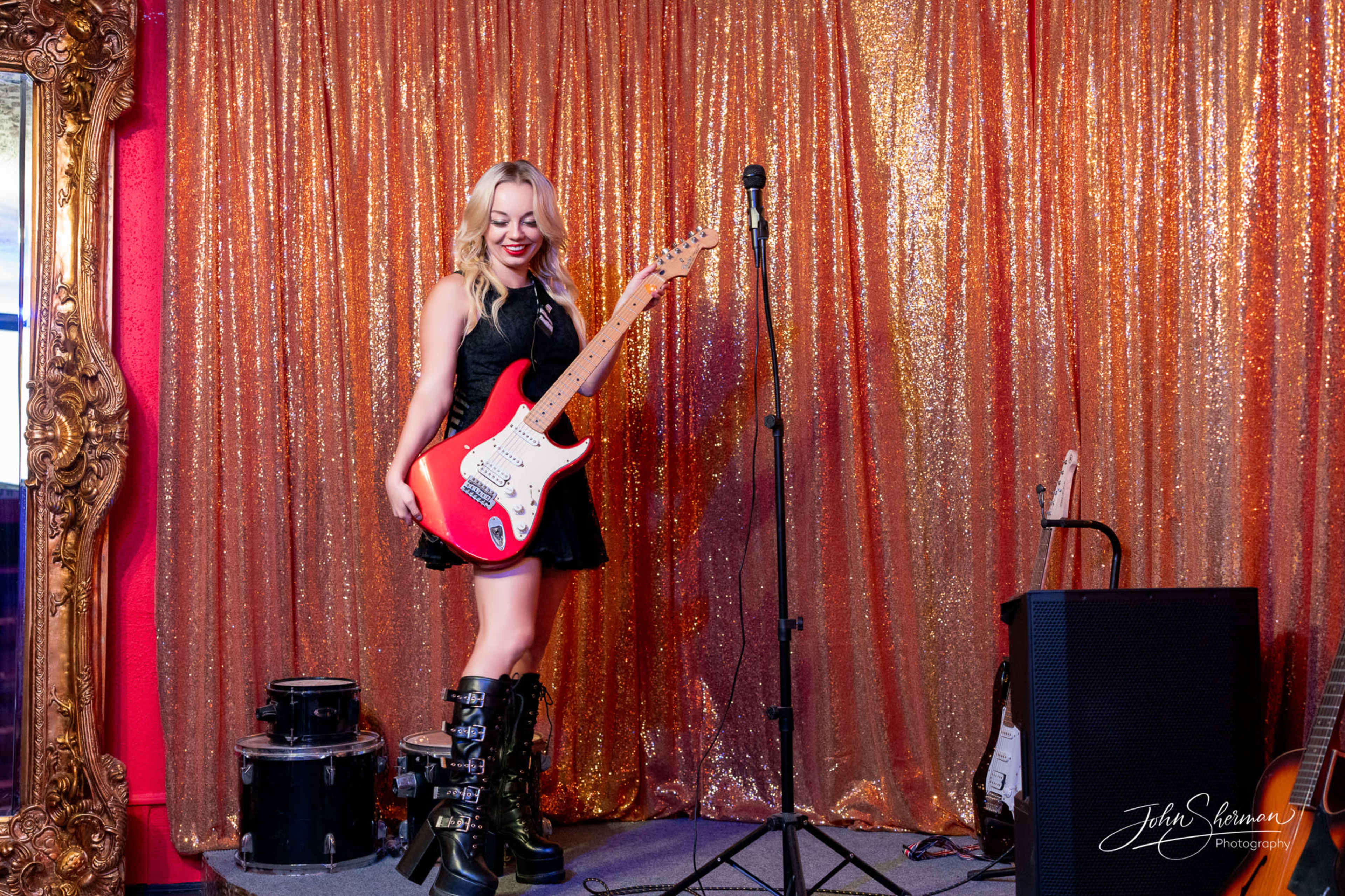 A woman in a black dress stands holding a red electric guitar in front of a sequined backdrop.