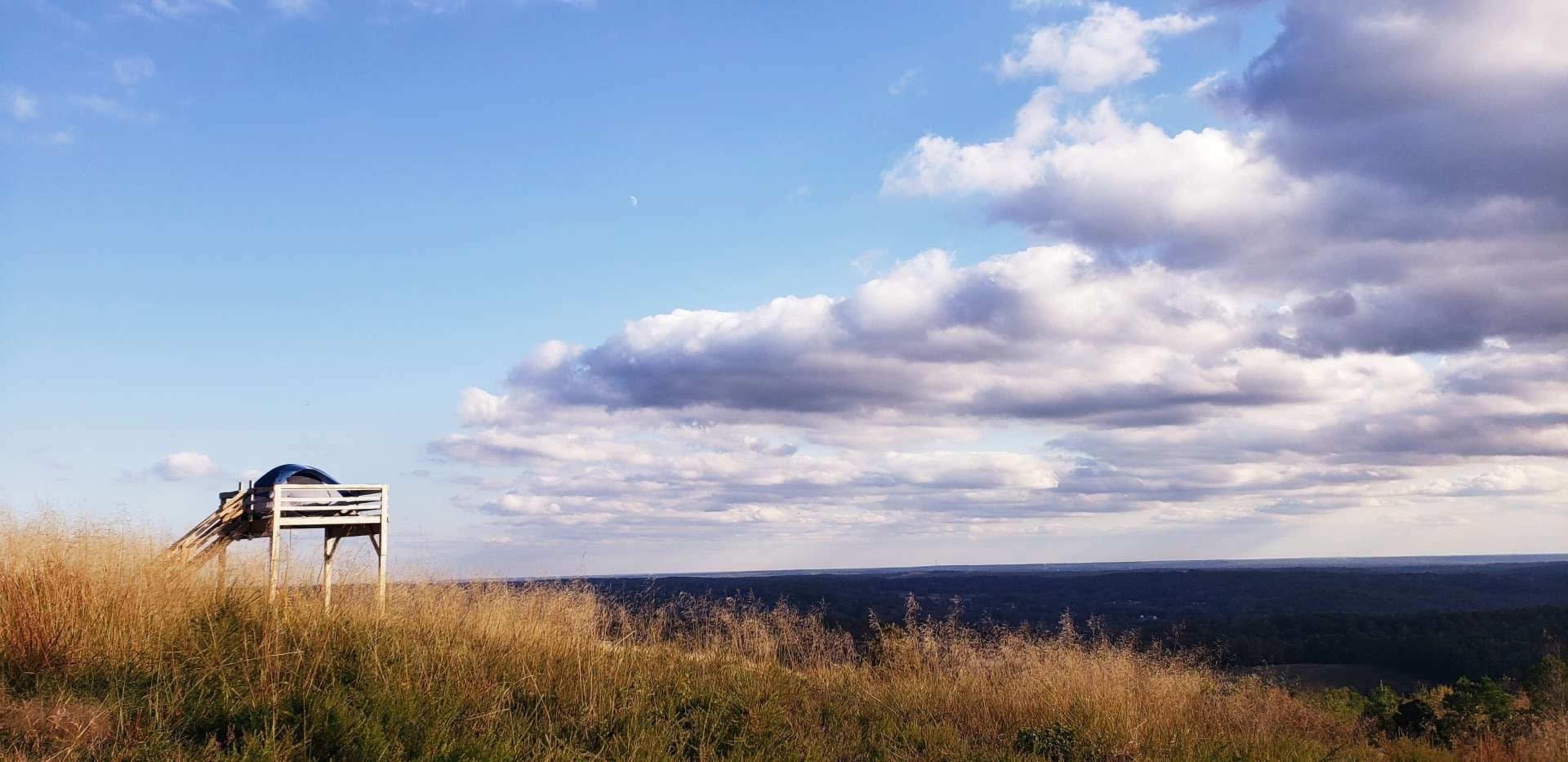 A wooden bench stands on a grassy hilltop overlooking a valley under a partly cloudy sky.