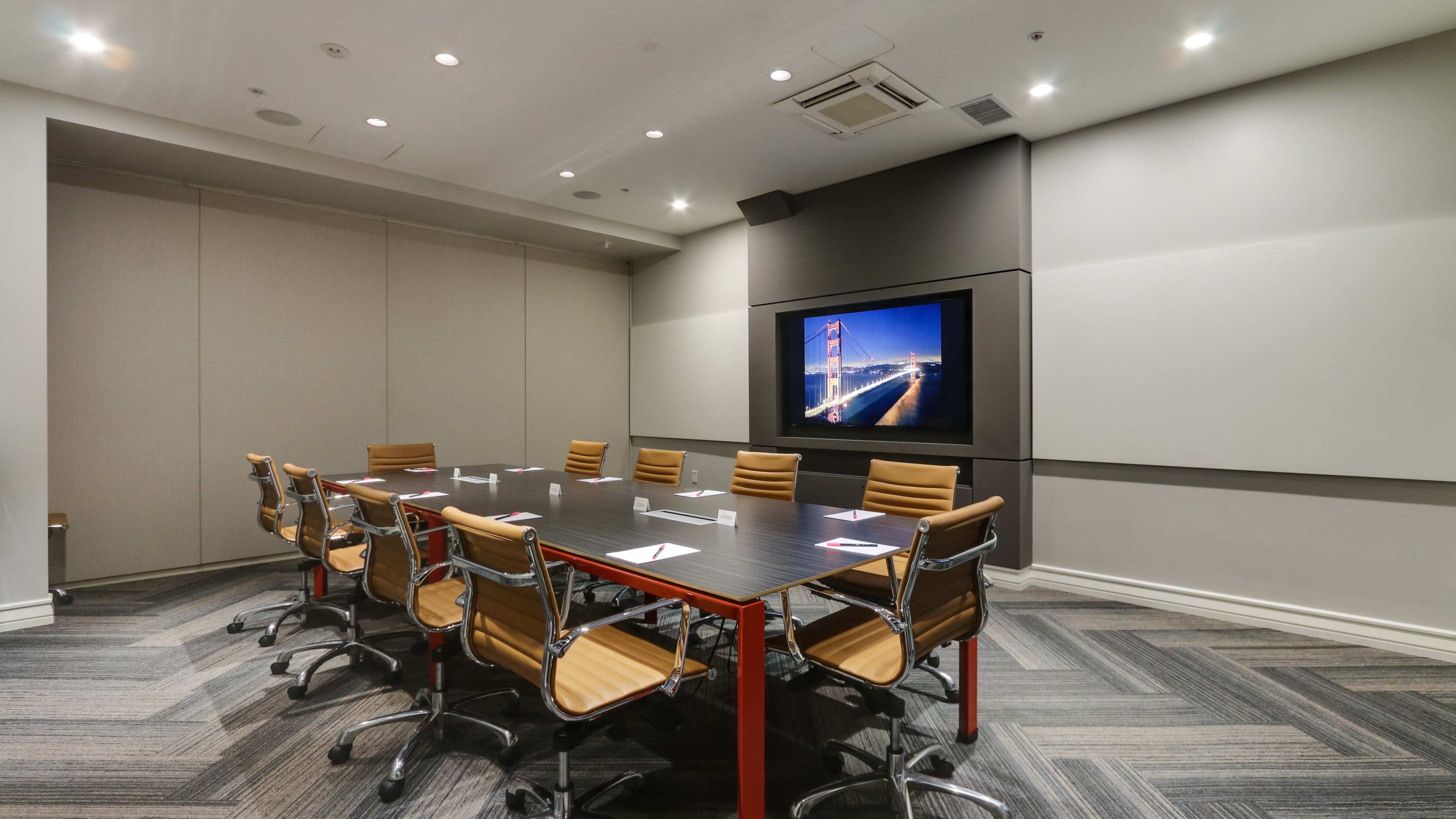 A conference room features a large table surrounded by ten chairs, with a wall-mounted screen displaying an image of the Golden Gate Bridge.