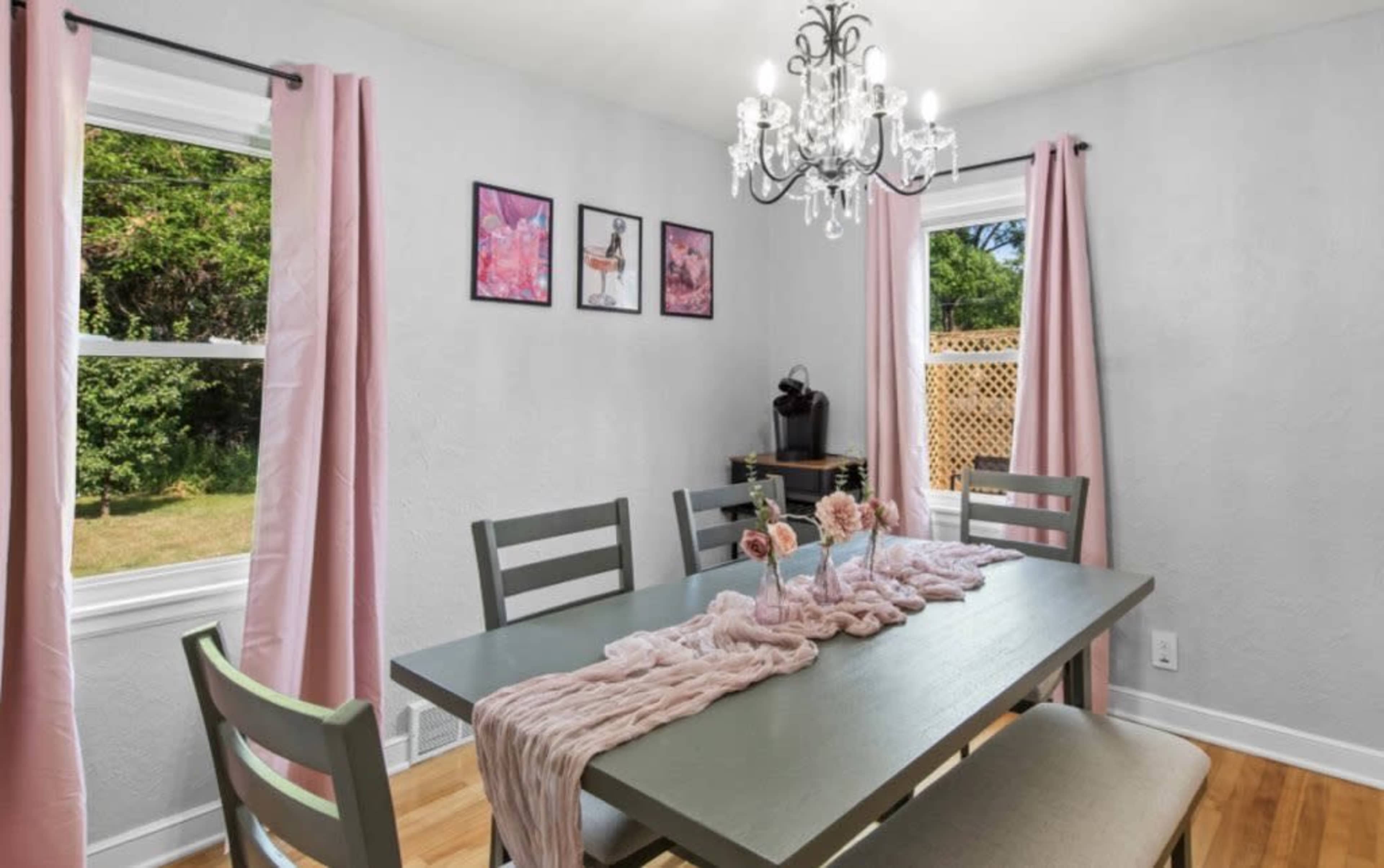 A dining room features a grey table surrounded by wooden chairs, with pink curtains framing two windows and a chandelier above.