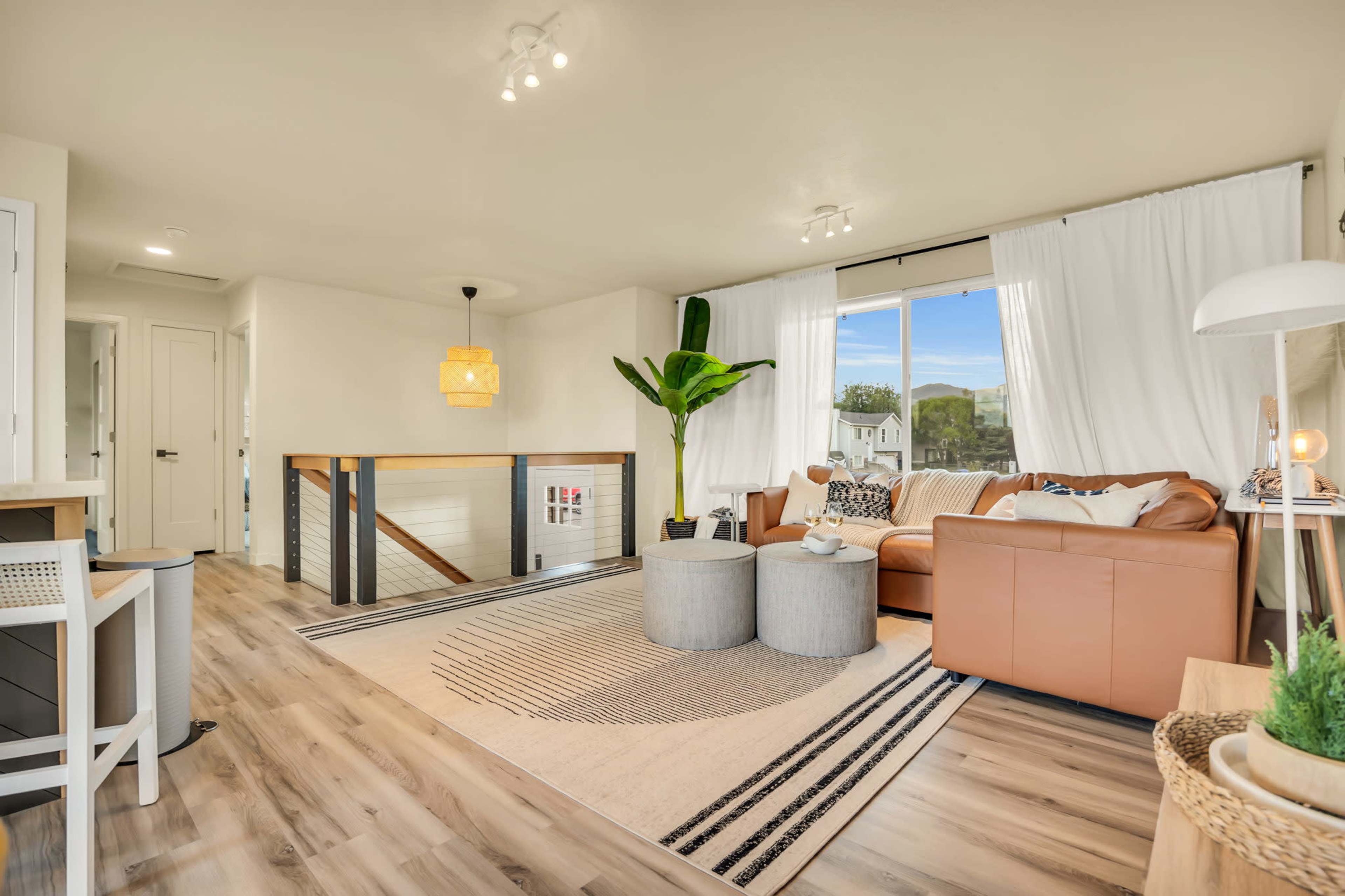 A cozy living room features a brown leather sofa, a large window with white curtains, and two round side tables on a patterned rug.