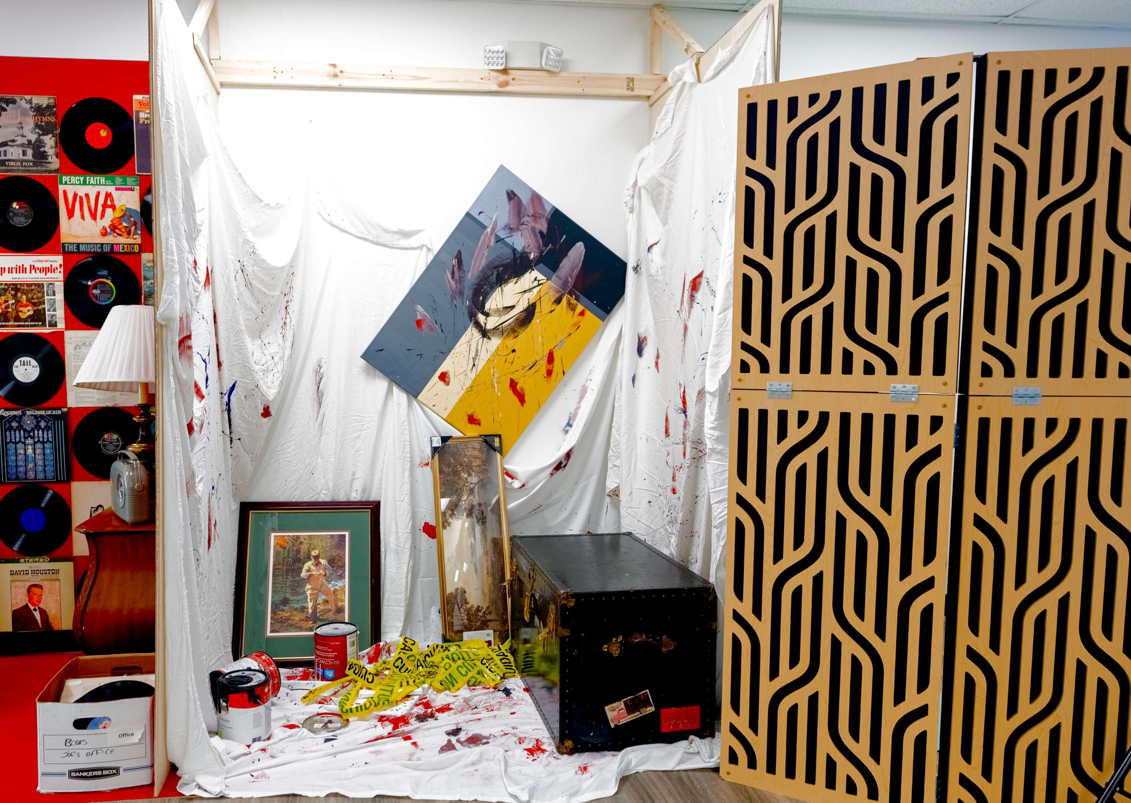 The image shows a cluttered corner of a room featuring a triangular piece of artwork with splashes of red, a framed picture, a black chest, and various items including paint cans and a lamp, all set against a backdrop of white fabric and patterned wooden panels.