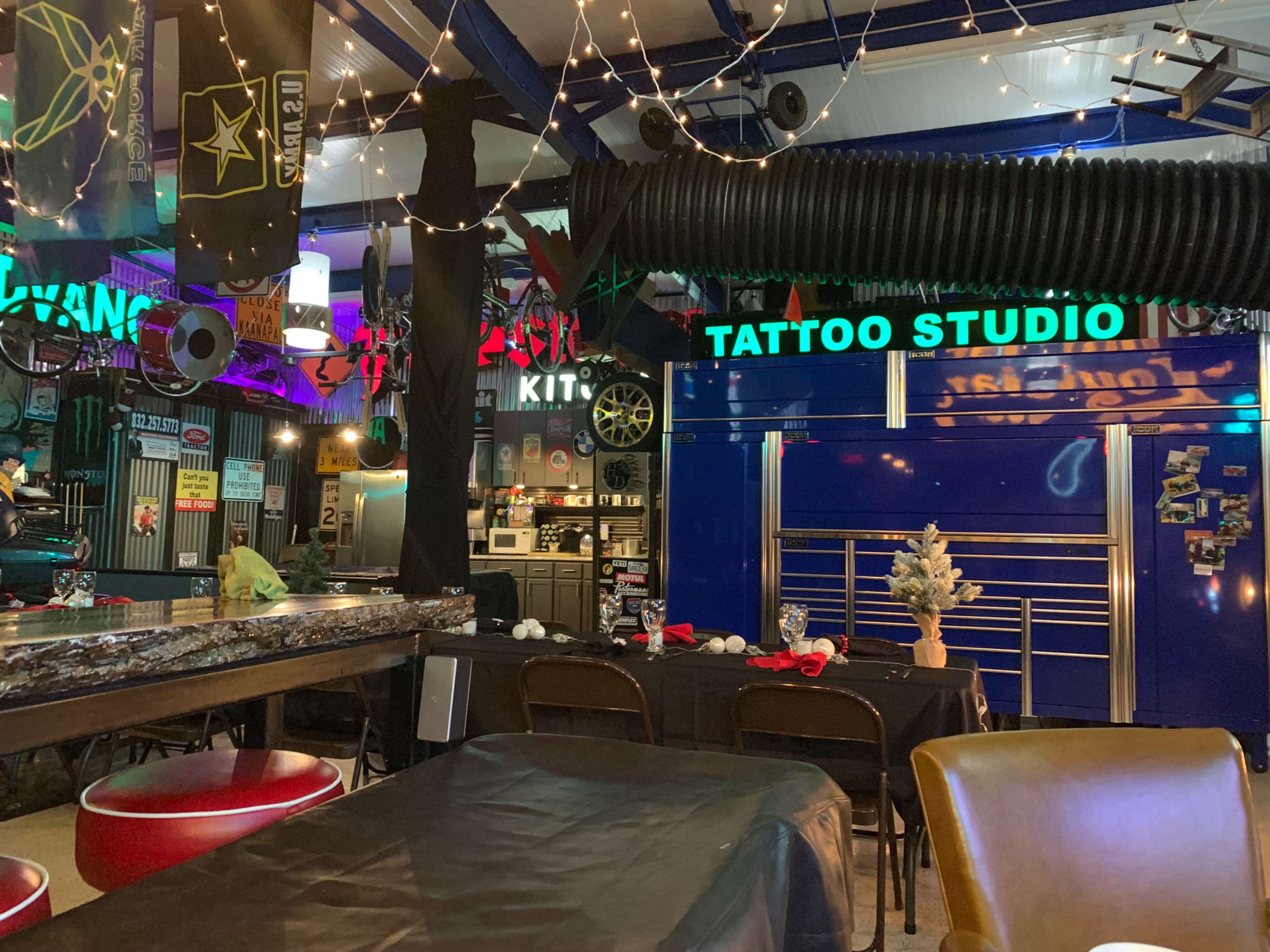 An indoor space featuring a bar area, a tattoo studio sign, and various colorful decorations, with tables set for dining.