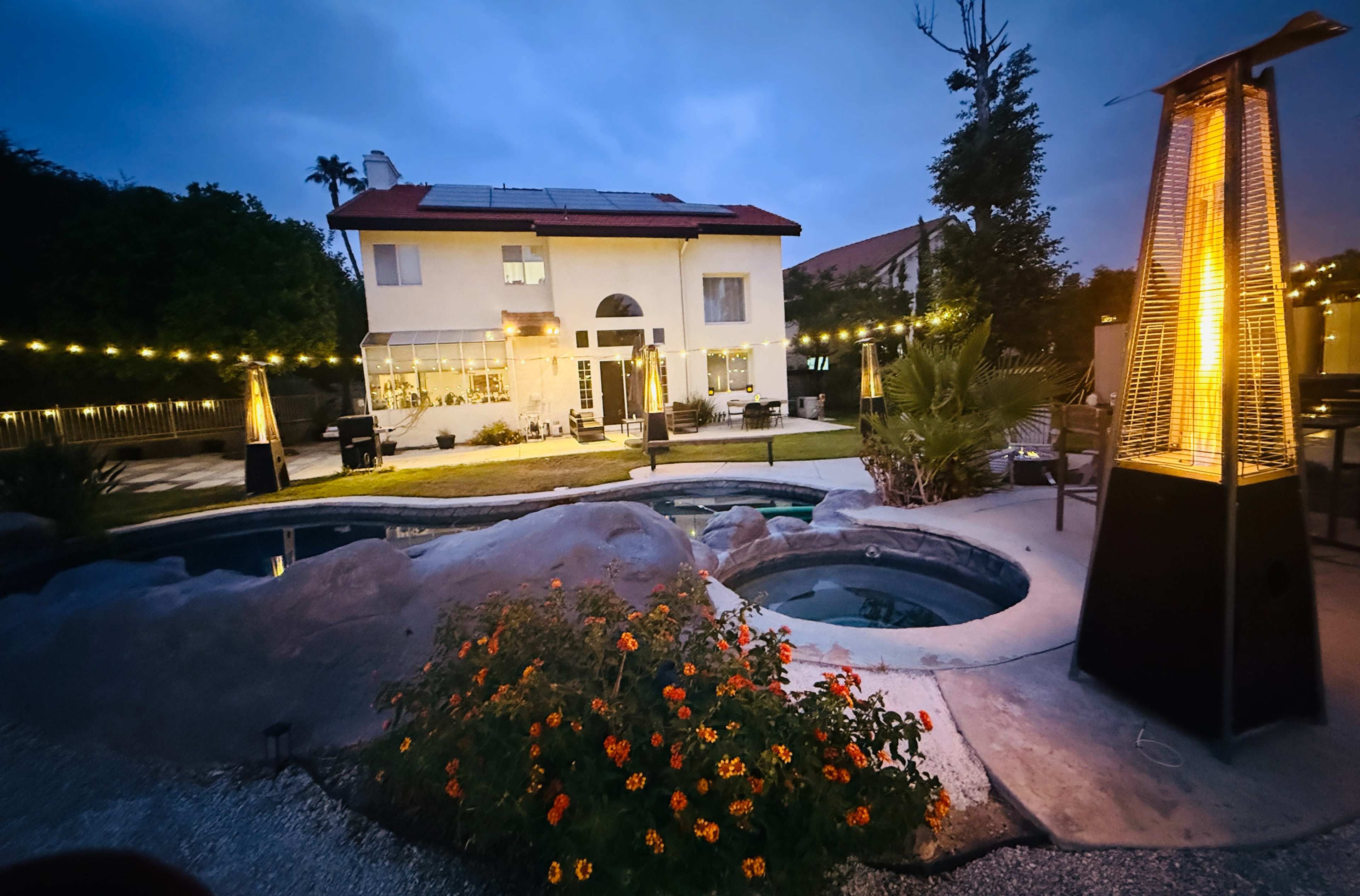 The image shows a two-story house with solar panels, a pool, and landscaped gardens illuminated by evening lights.