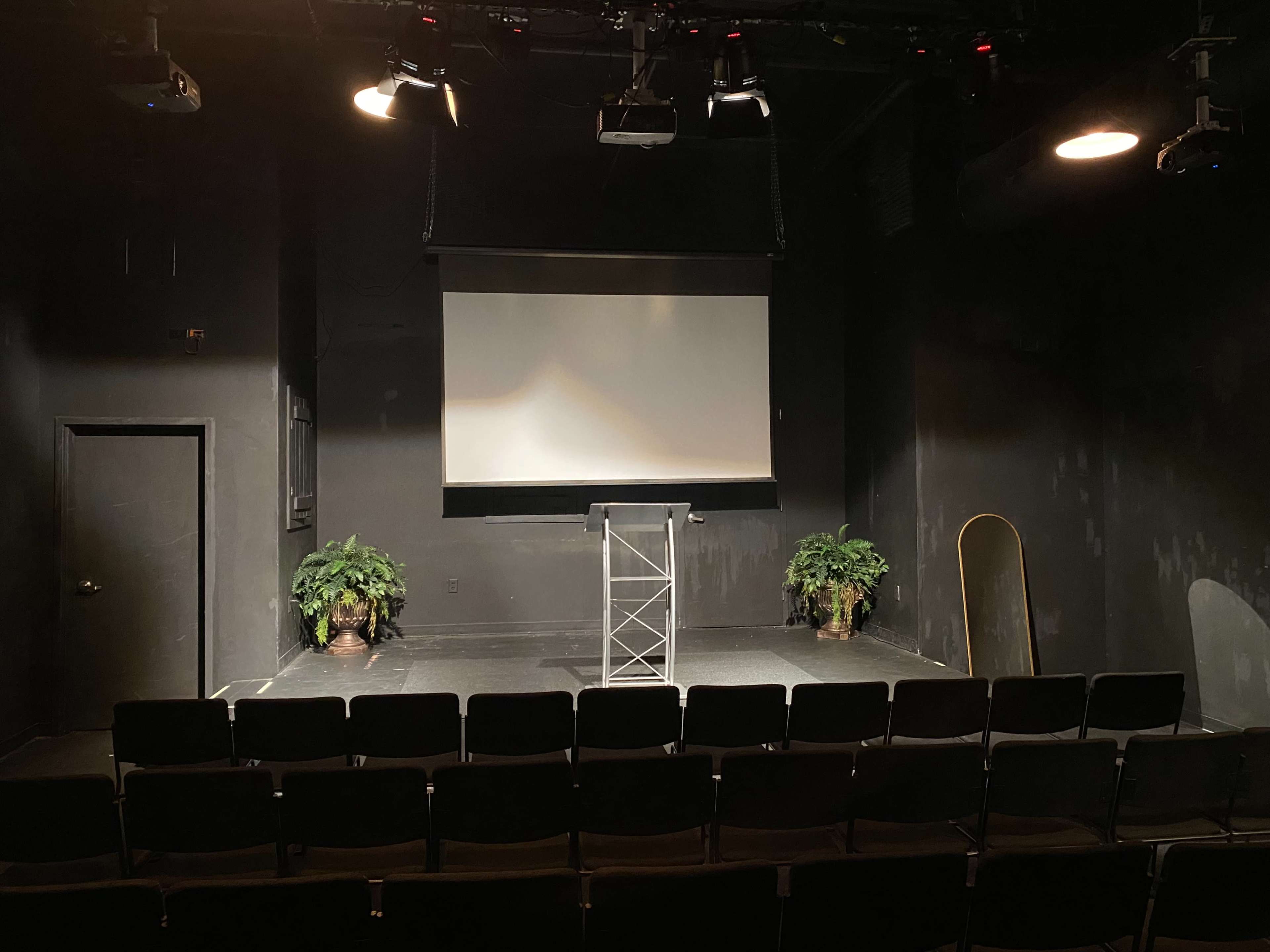 A dimly lit stage with a podium, a projector screen, two potted plants, and rows of black chairs facing the front.