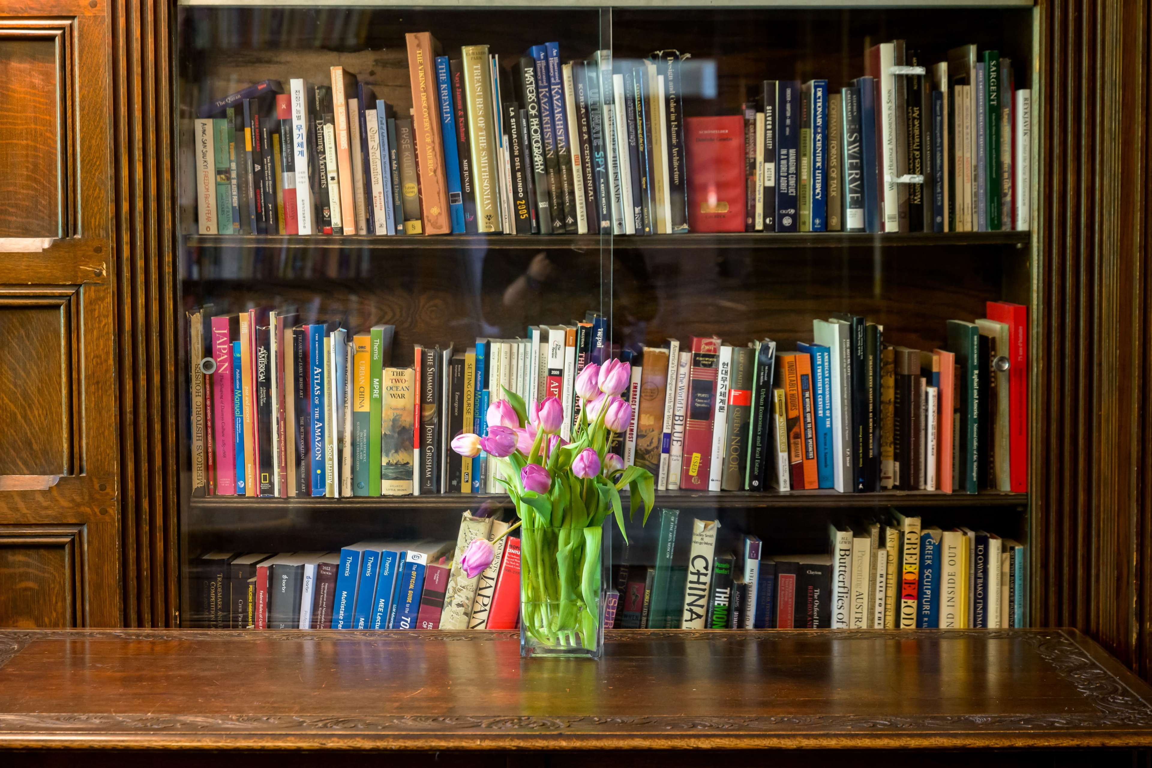 A wooden table displays a vase of pink tulips in front of a bookshelf filled with various books.