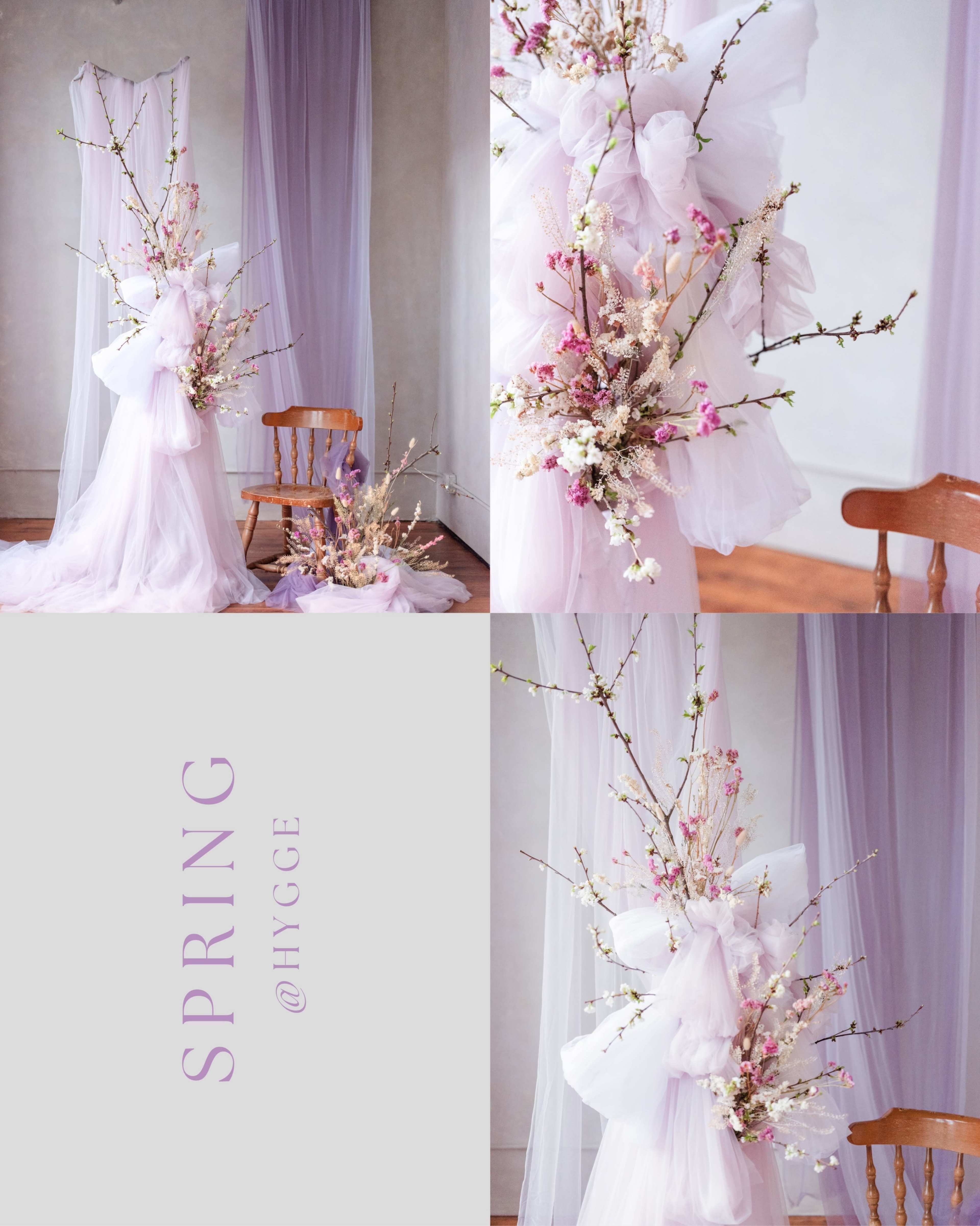 The image features a decorative arrangement of branches and flowers intertwined with soft, flowing fabric in pastel colors, set against a backdrop of draped sheer curtains and wooden chairs.