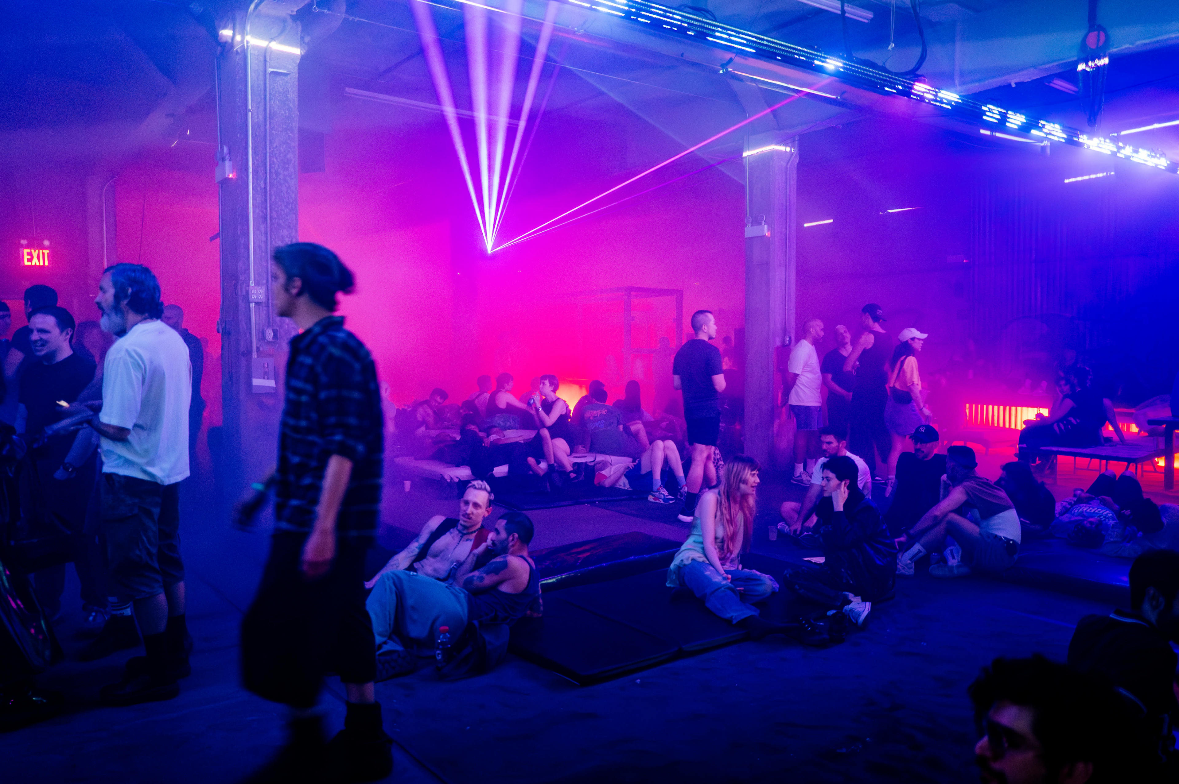 A vibrant indoor event space is filled with people socializing and lounging under colorful laser lights and a haze of fog.