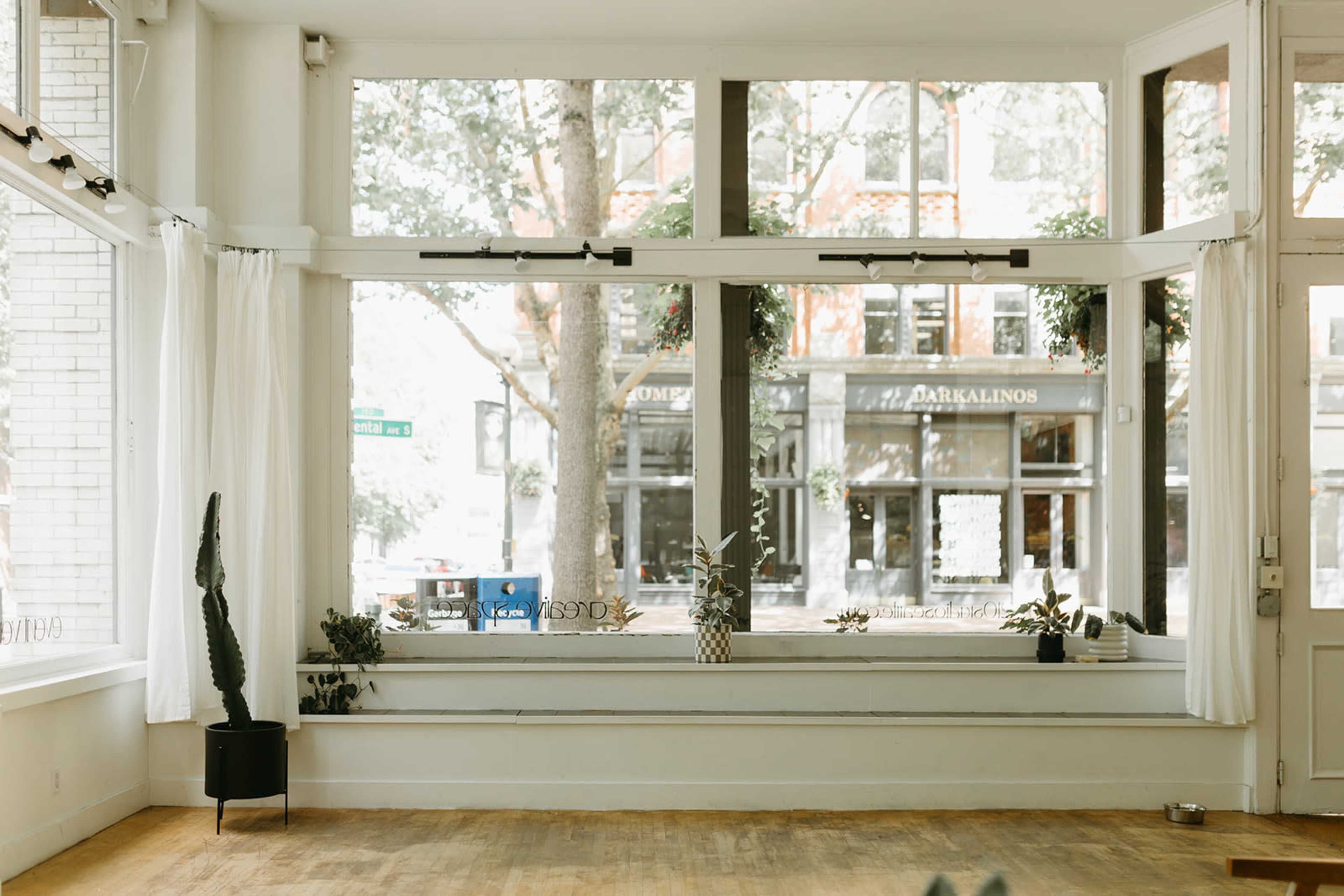 The image shows a spacious, bright interior with large windows facing a city street, featuring simple decor including plants and a light wooden floor.