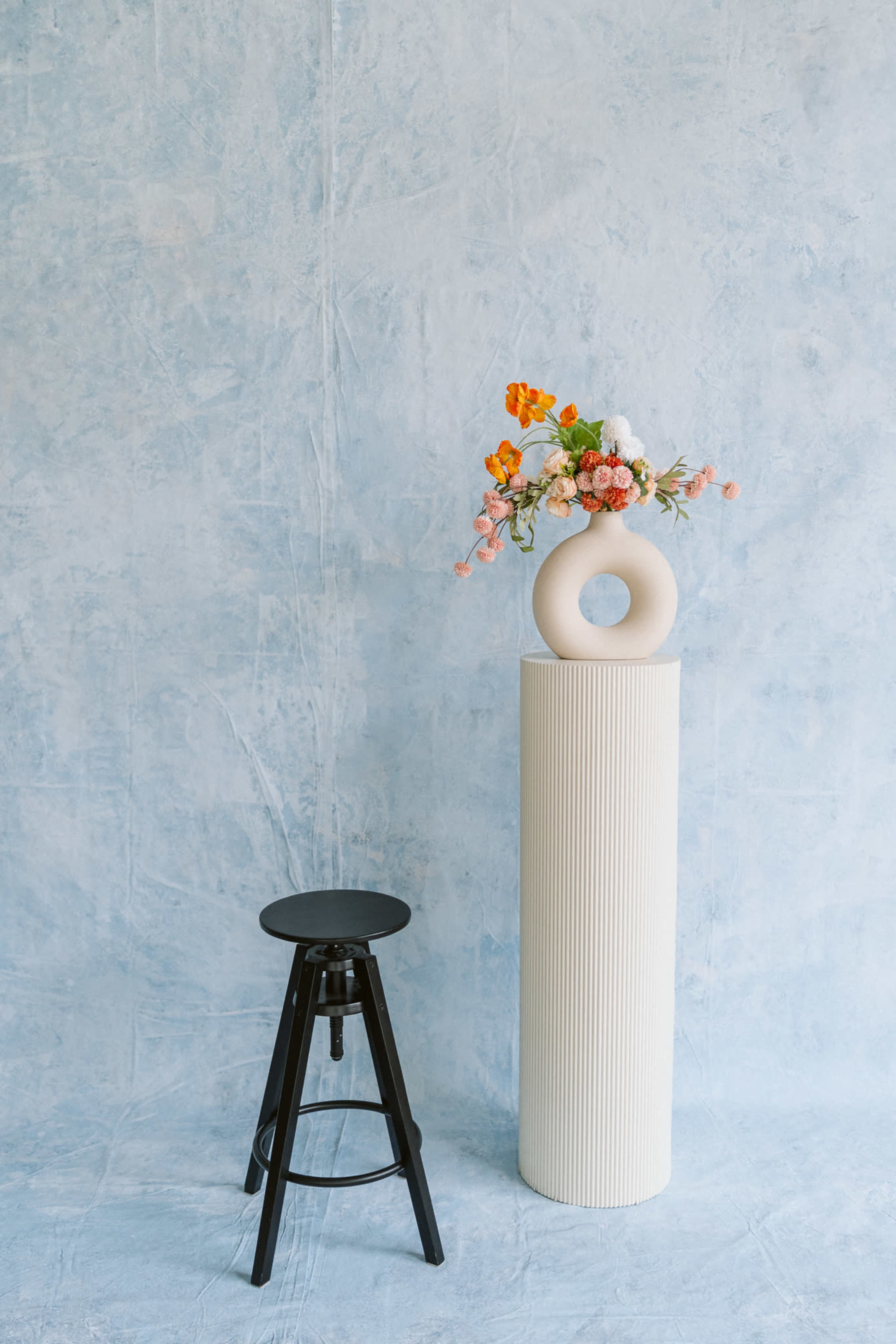 A white column with a circular vase of flowers stands next to a black stool against a light blue background.