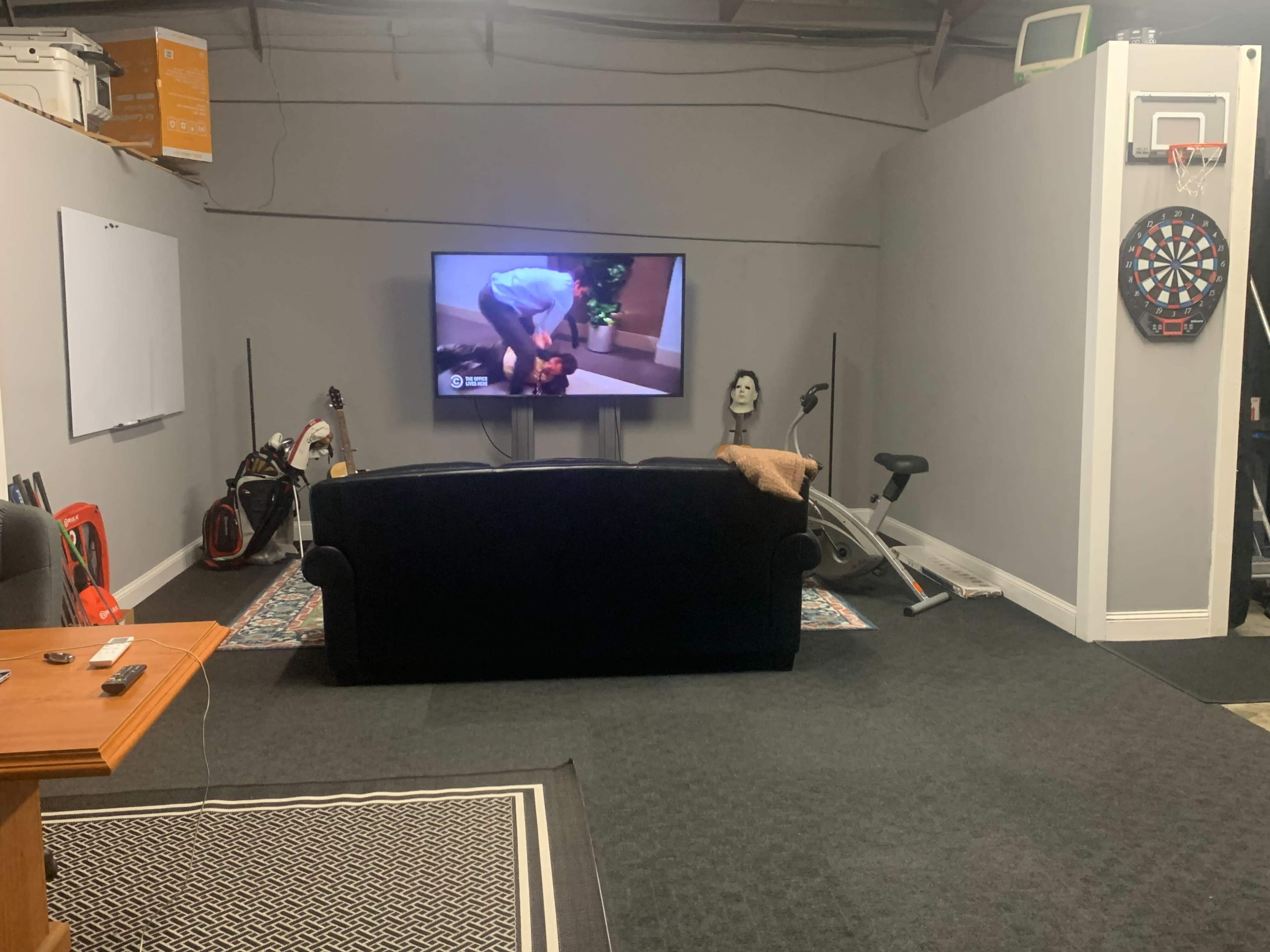 A garage-like room with a black couch facing a television, along with a dartboard, exercise bike, and various sports equipment.