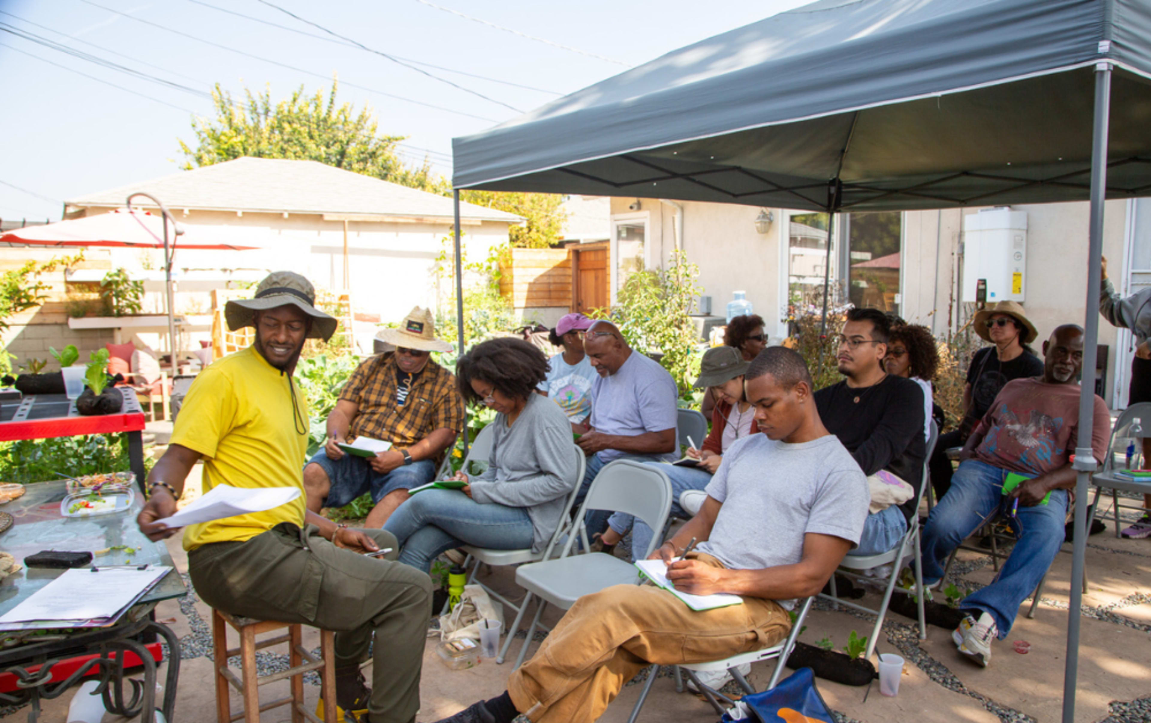 A group of people sits beneath a canopy in a garden setting, engaged in a learning or discussion activity.
