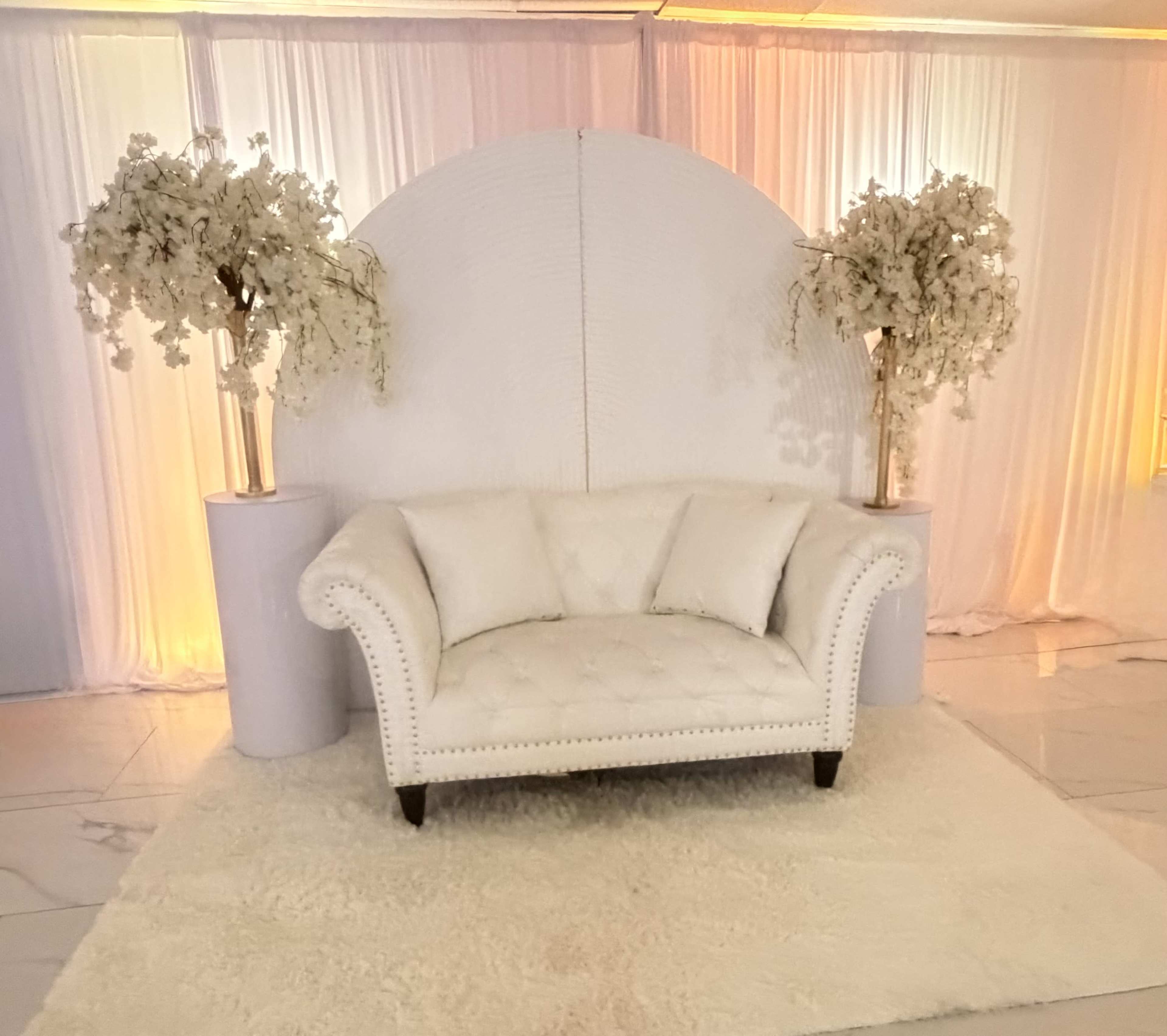 A white, tufted couch is positioned in front of a circular backdrop adorned with white floral arrangements on either side, set against a softly lit draped curtain.