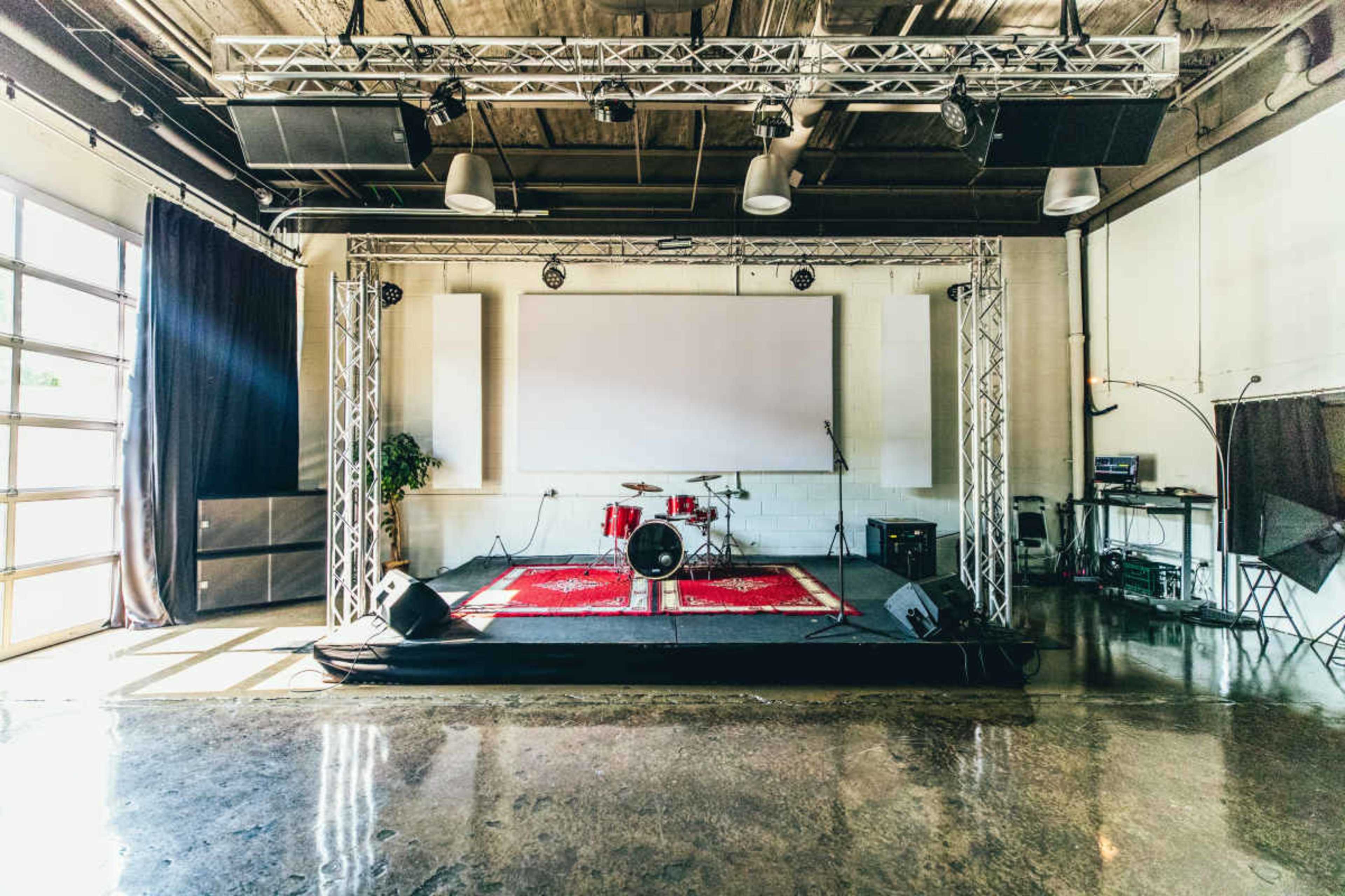 The image shows a performance stage with drum equipment, a projector screen, and sound equipment in a spacious room.