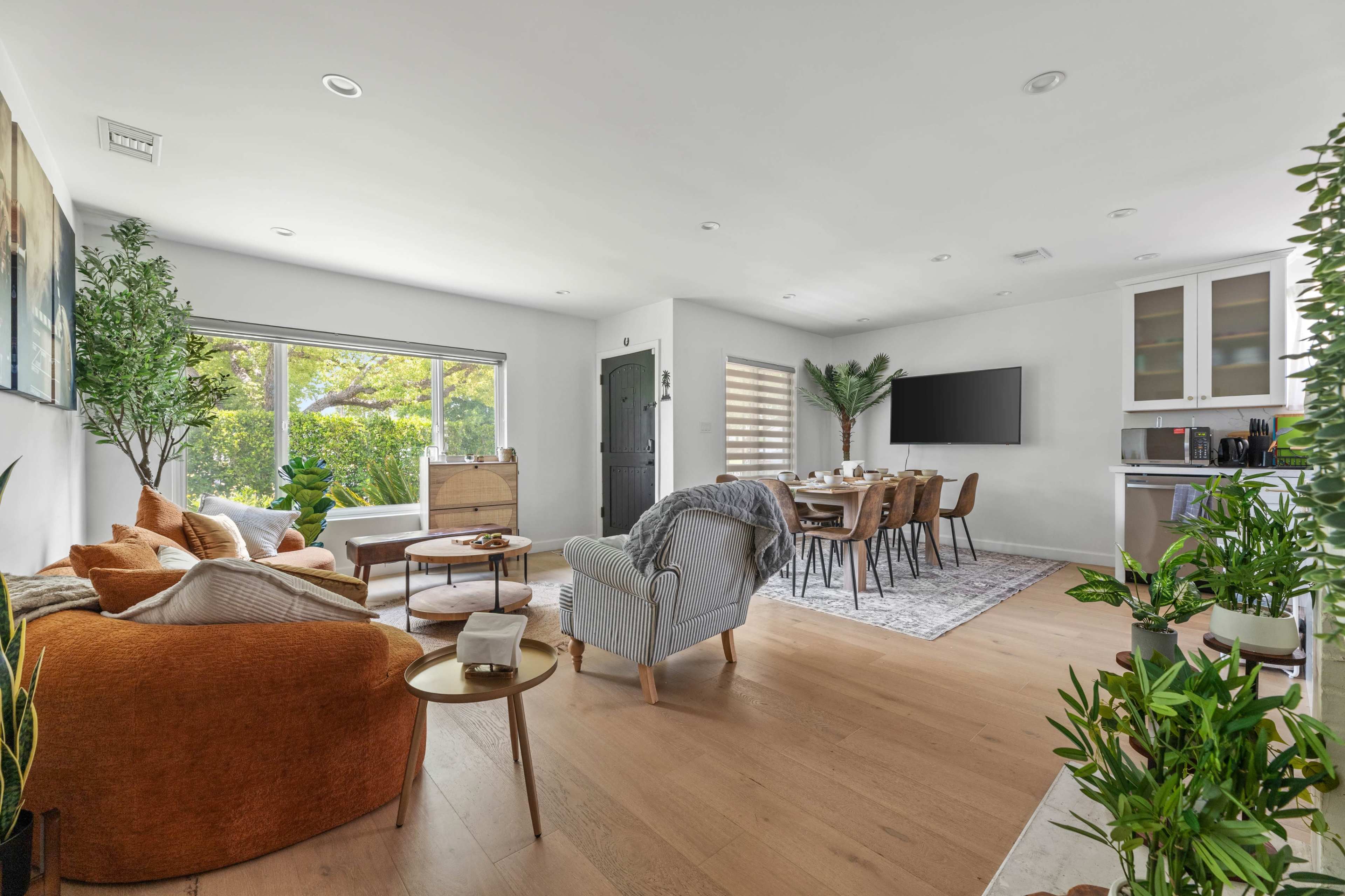 The image shows a modern living area with a cozy seating arrangement, a dining table for six, and large windows letting in natural light.