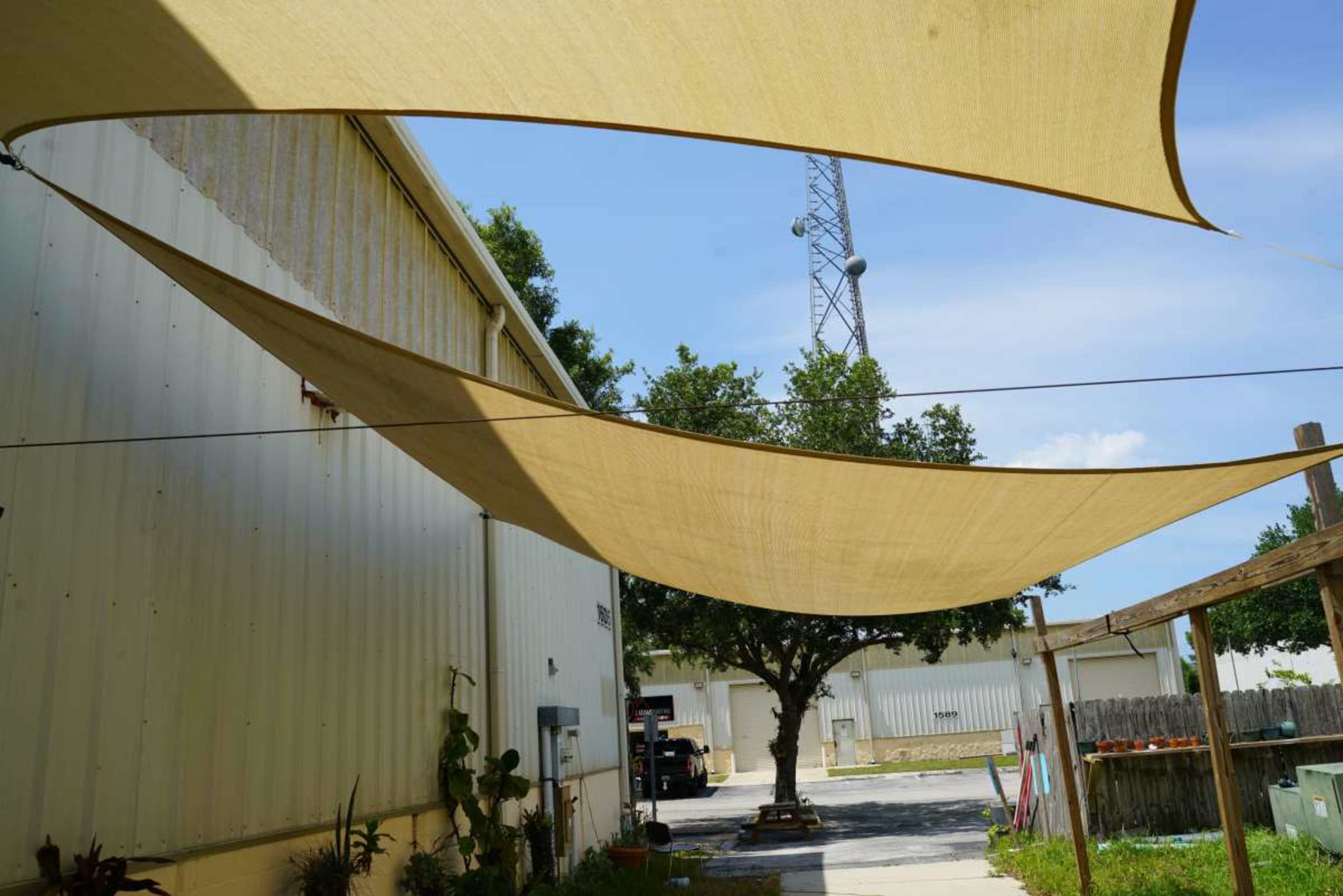 The image shows large fabric canopies stretched overhead in an outdoor area adjacent to a building, with a tree and a telecommunications tower visible in the background.