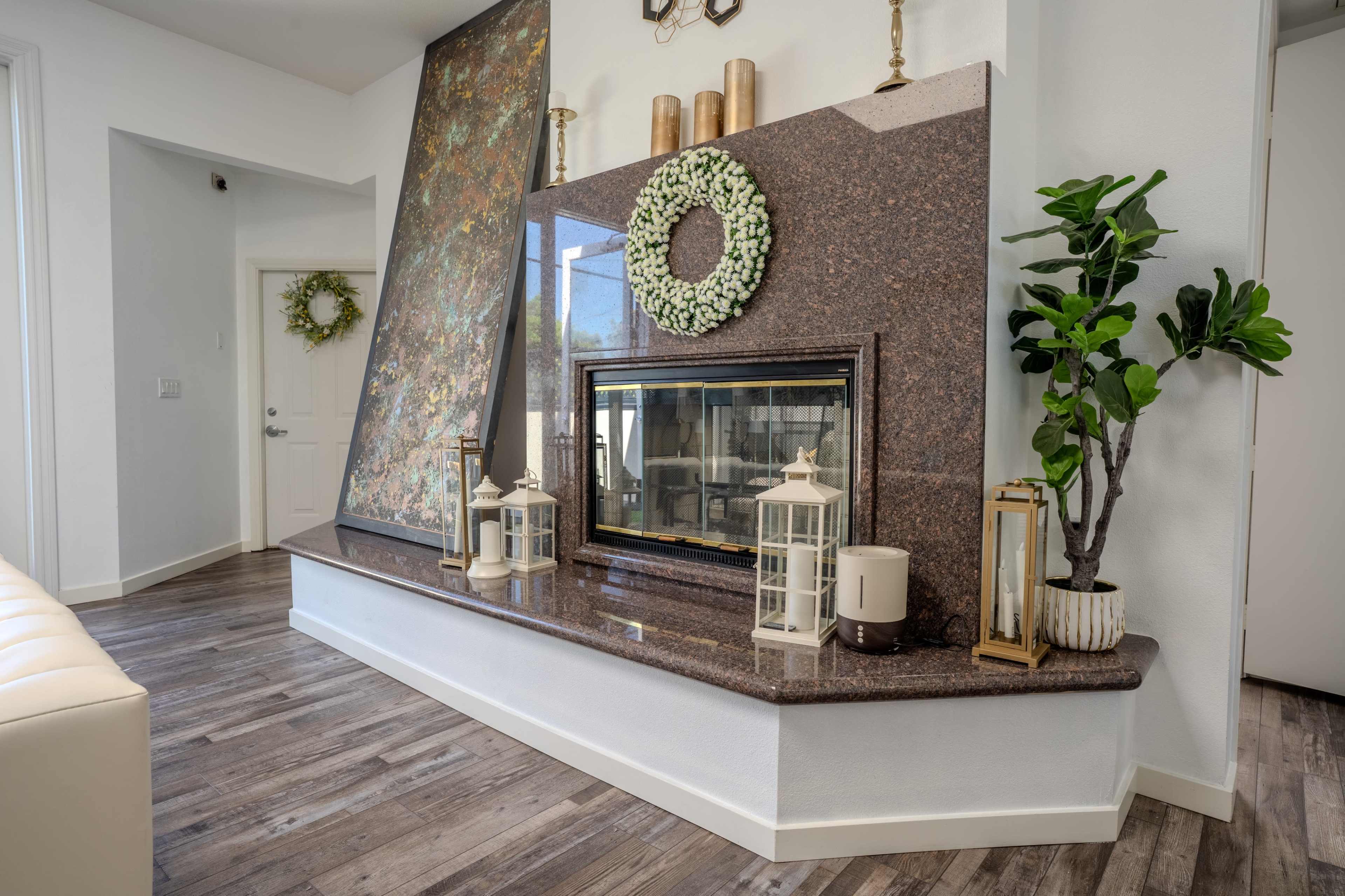 The image shows a modern living room with a granite fireplace, decorative plants, and lanterns arranged on a wooden floor.