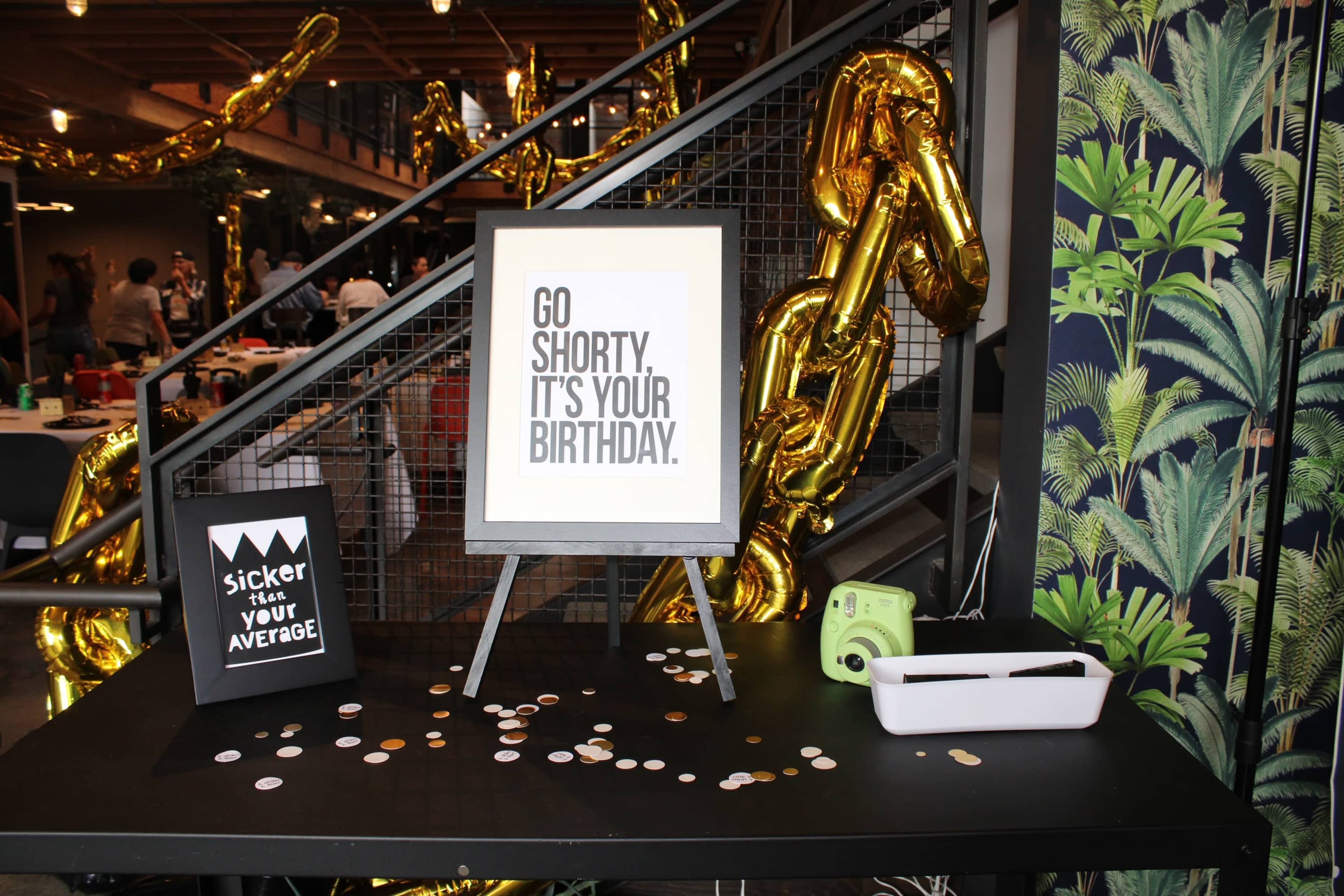 A decorated table features a sign reading "Go Shorty, It's Your Birthday" alongside a smaller sign that says "Sicker than Your Average," with gold balloon decorations in the background.