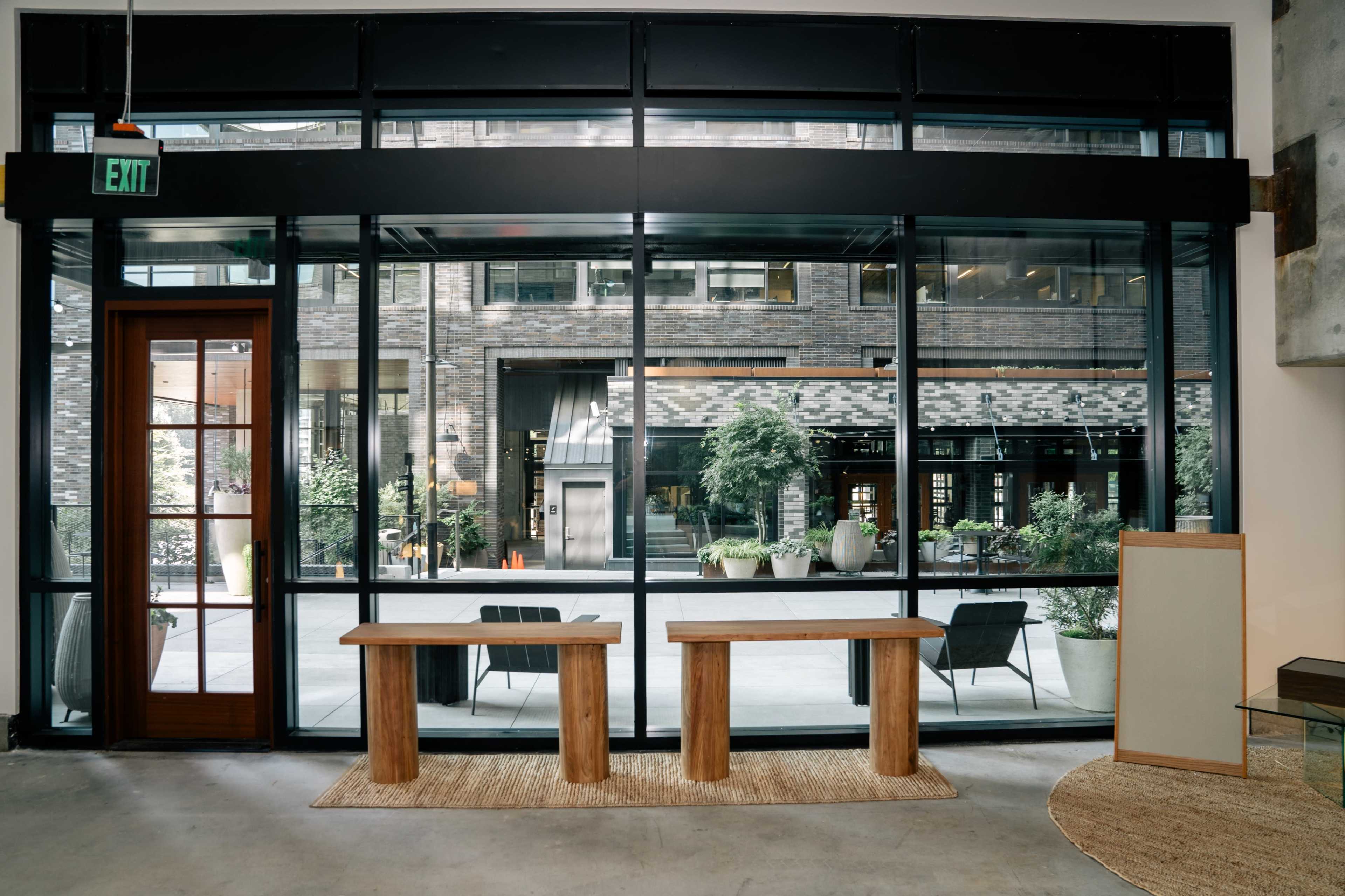 The image shows a modern interior space with large glass windows overlooking a courtyard, featuring two wooden tables and plants in pots.