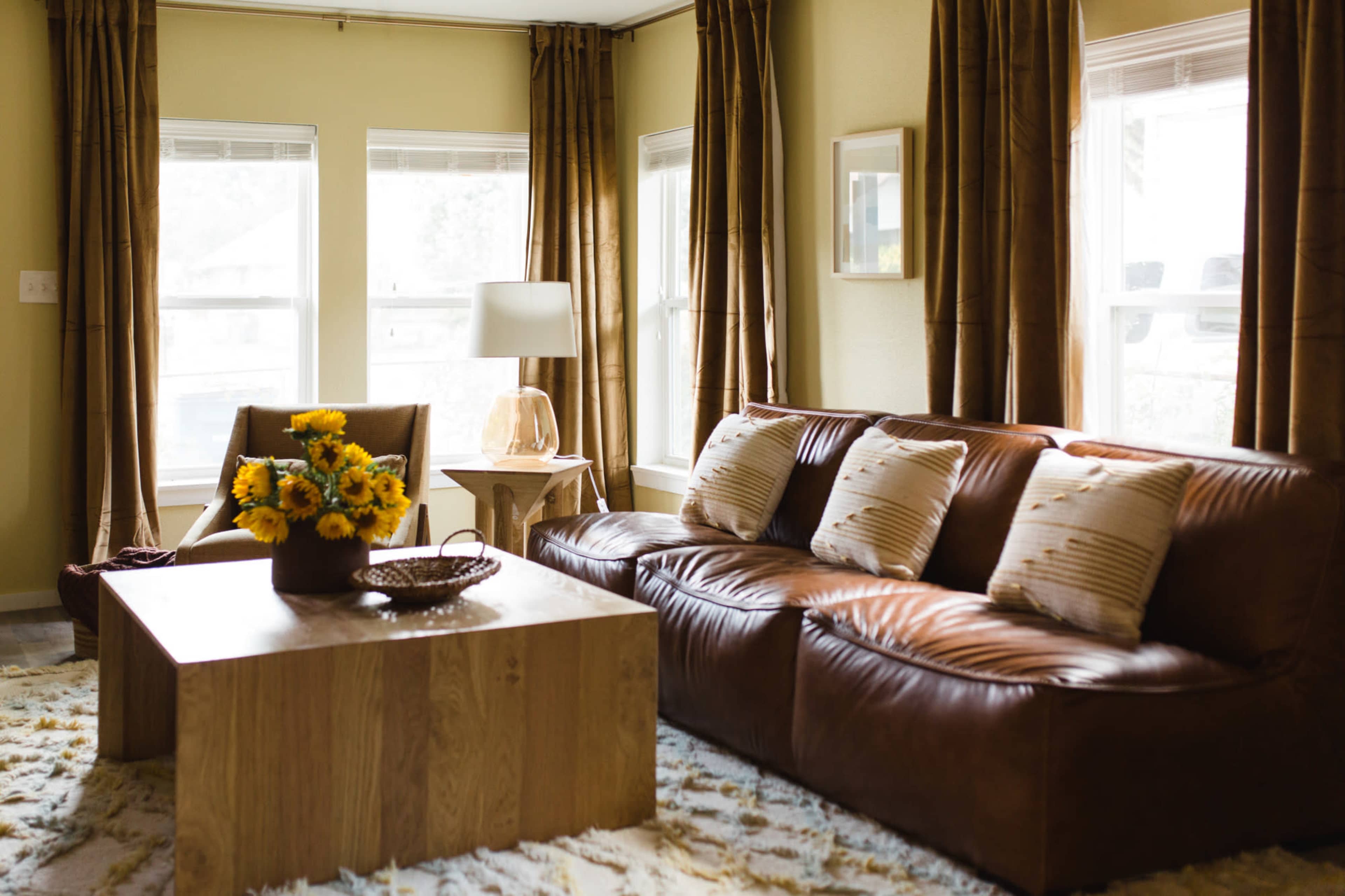 A living room features a brown leather sofa, a wooden coffee table, and a bouquet of sunflowers on the table, with sunlight streaming through the windows.