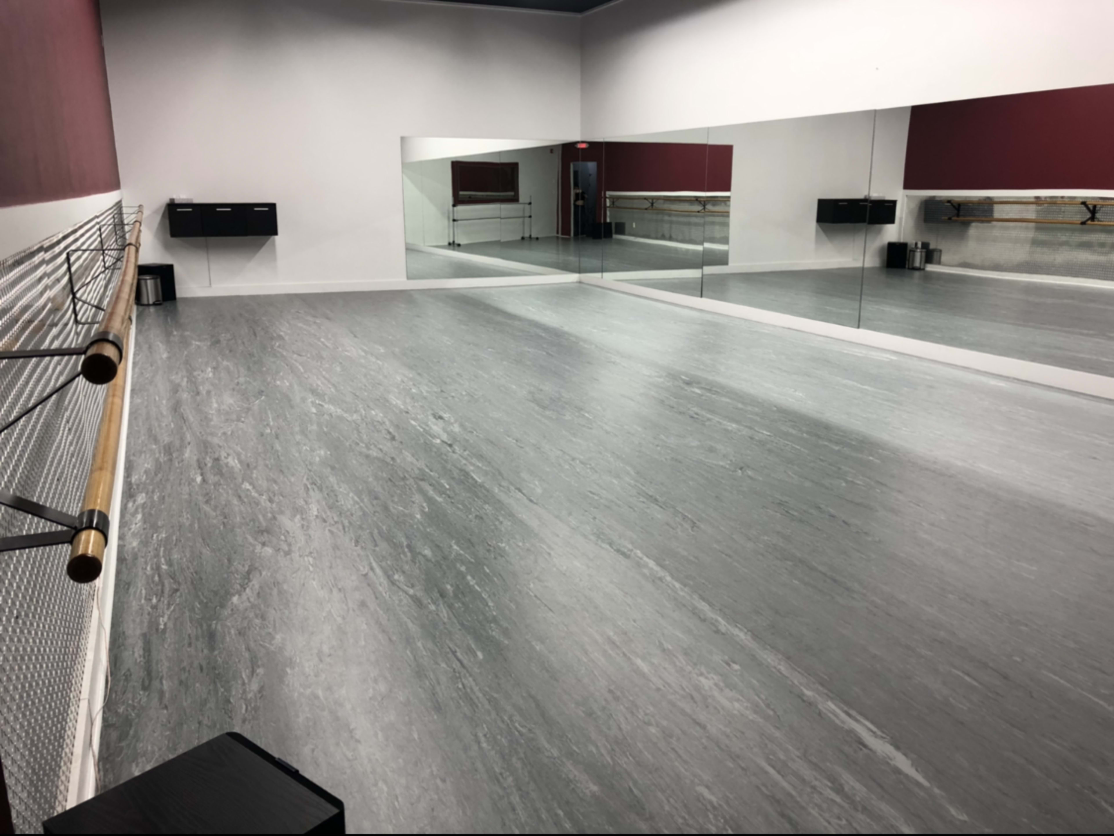 A spacious dance studio with a mirrored wall, wooden ballet bar, and gray flooring.