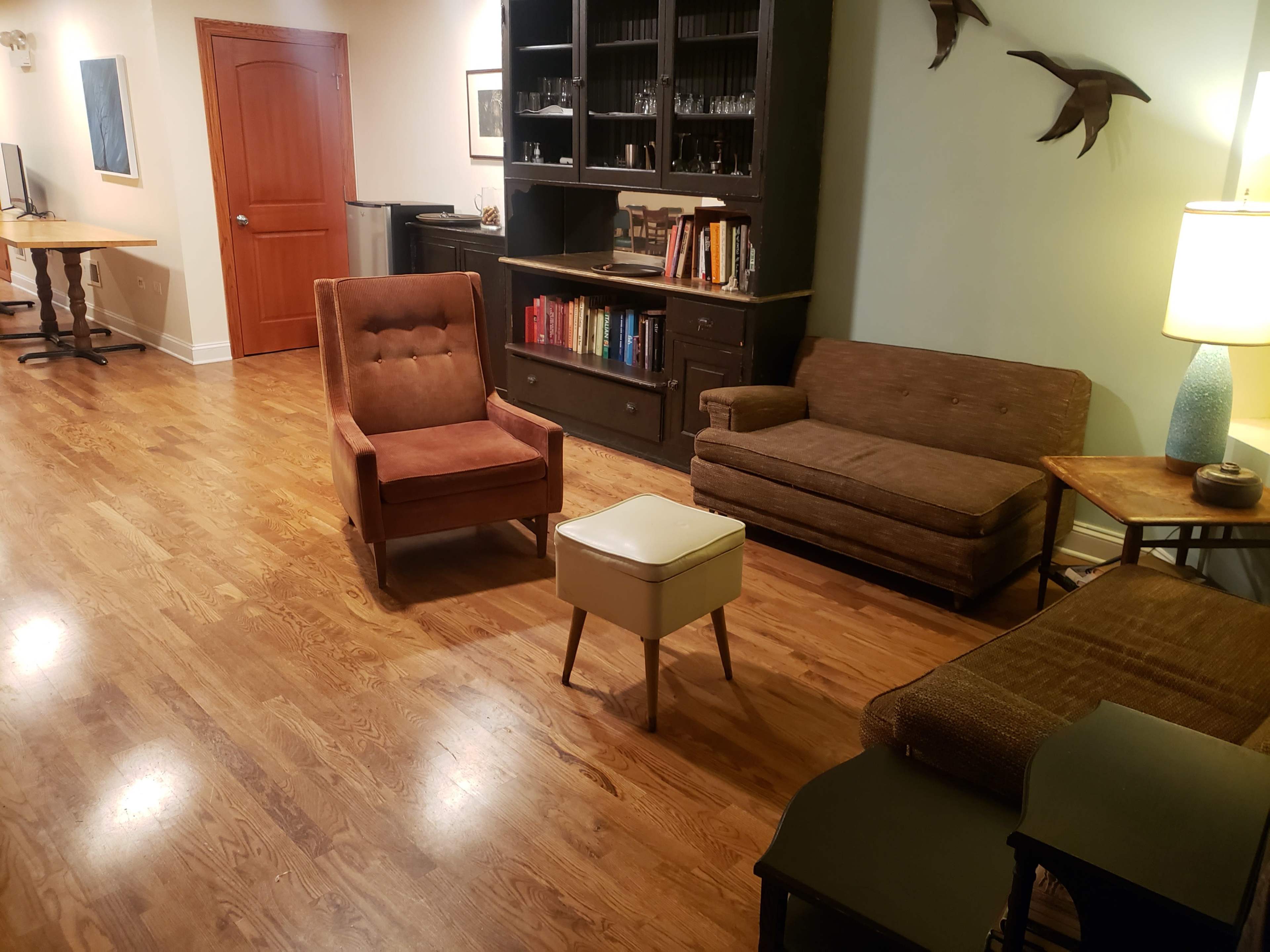 The room features hardwood flooring with a sofa, armchair, and coffee table arranged around a bookshelf and illuminated by a lamp.