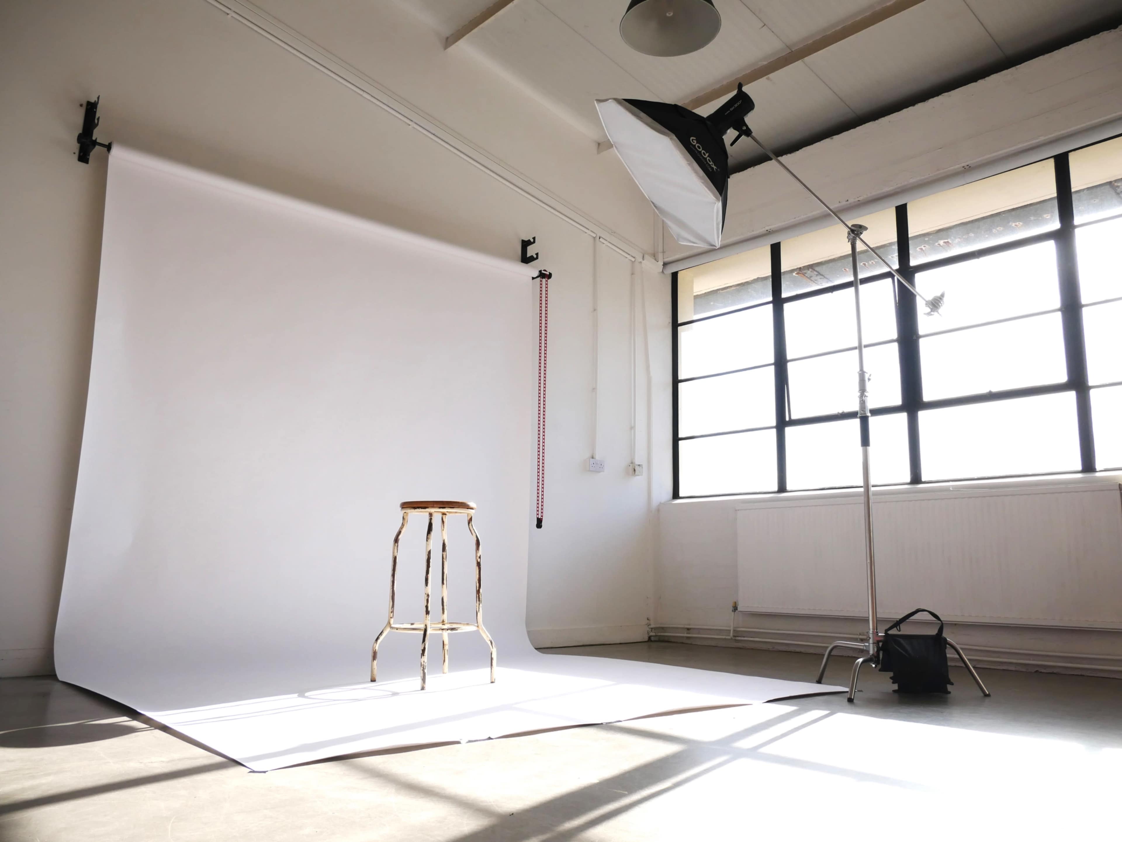 A well-lit photography studio features a white backdrop, a wooden stool, and a large softbox light setup near a large window.