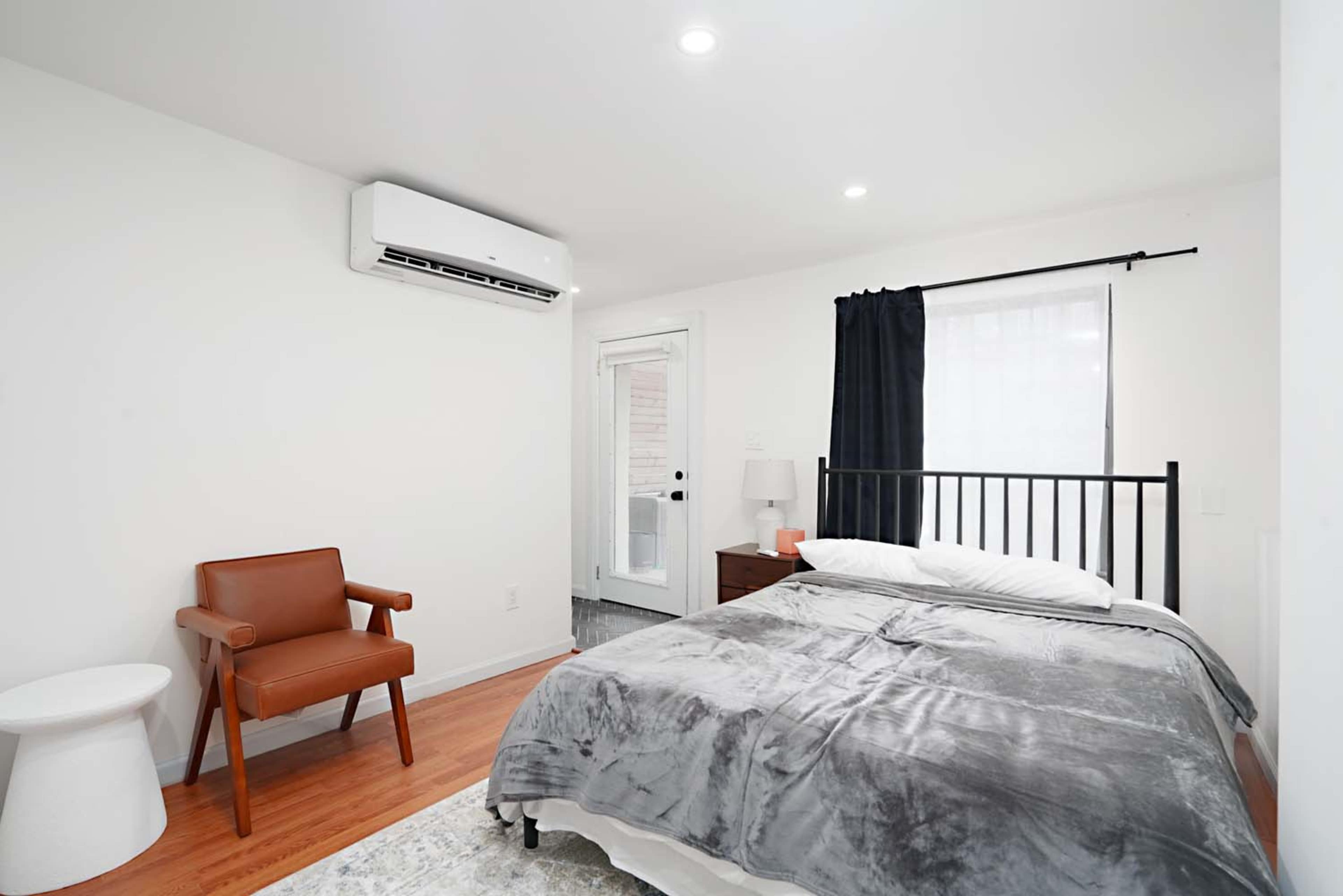 The image shows a modern bedroom with a bed, a chair, a side table, and a window covered by a curtain.