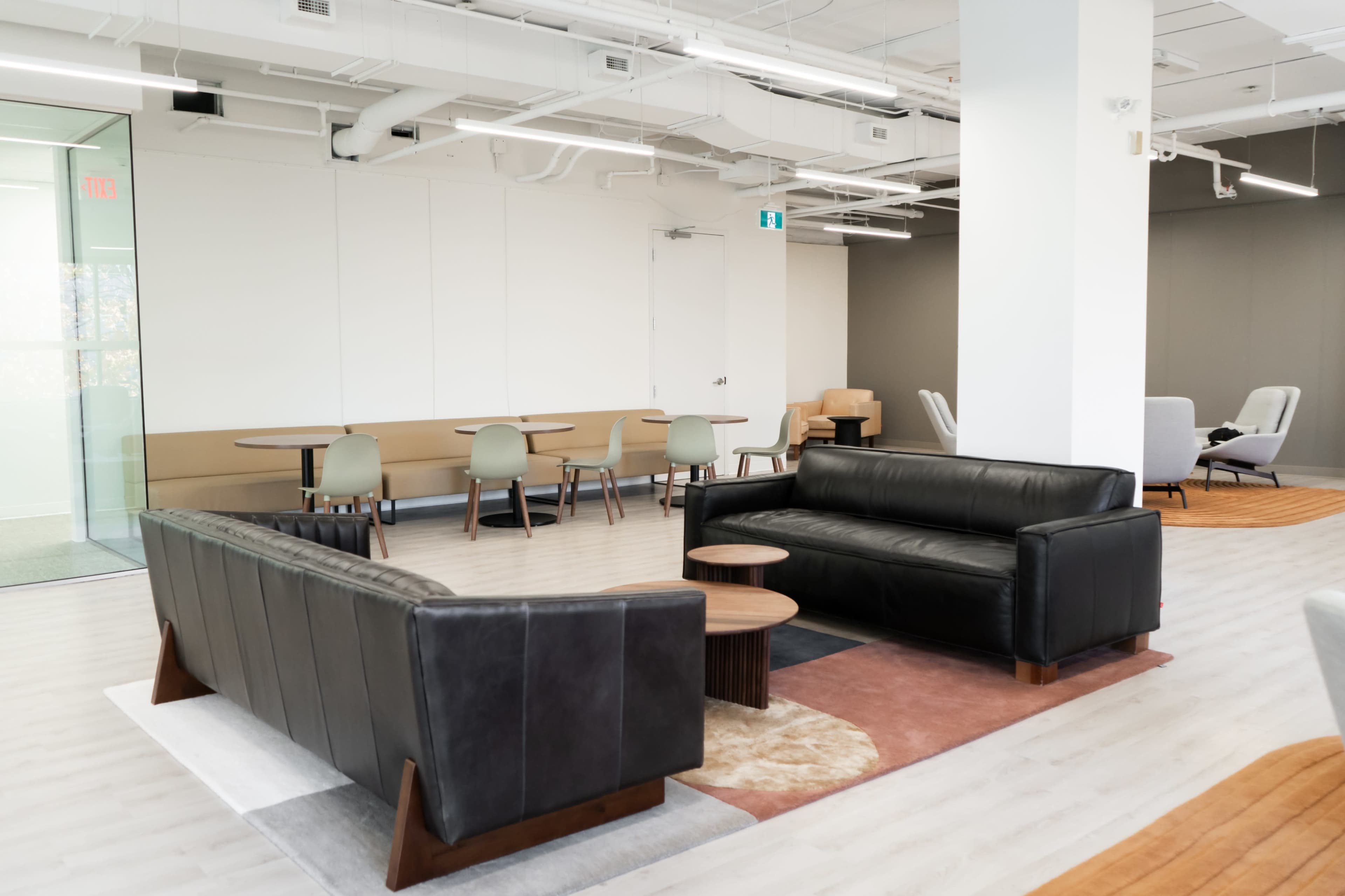 The image shows a modern office lounge area with black leather sofas, wooden coffee tables, and several chairs arranged around light-colored floors.