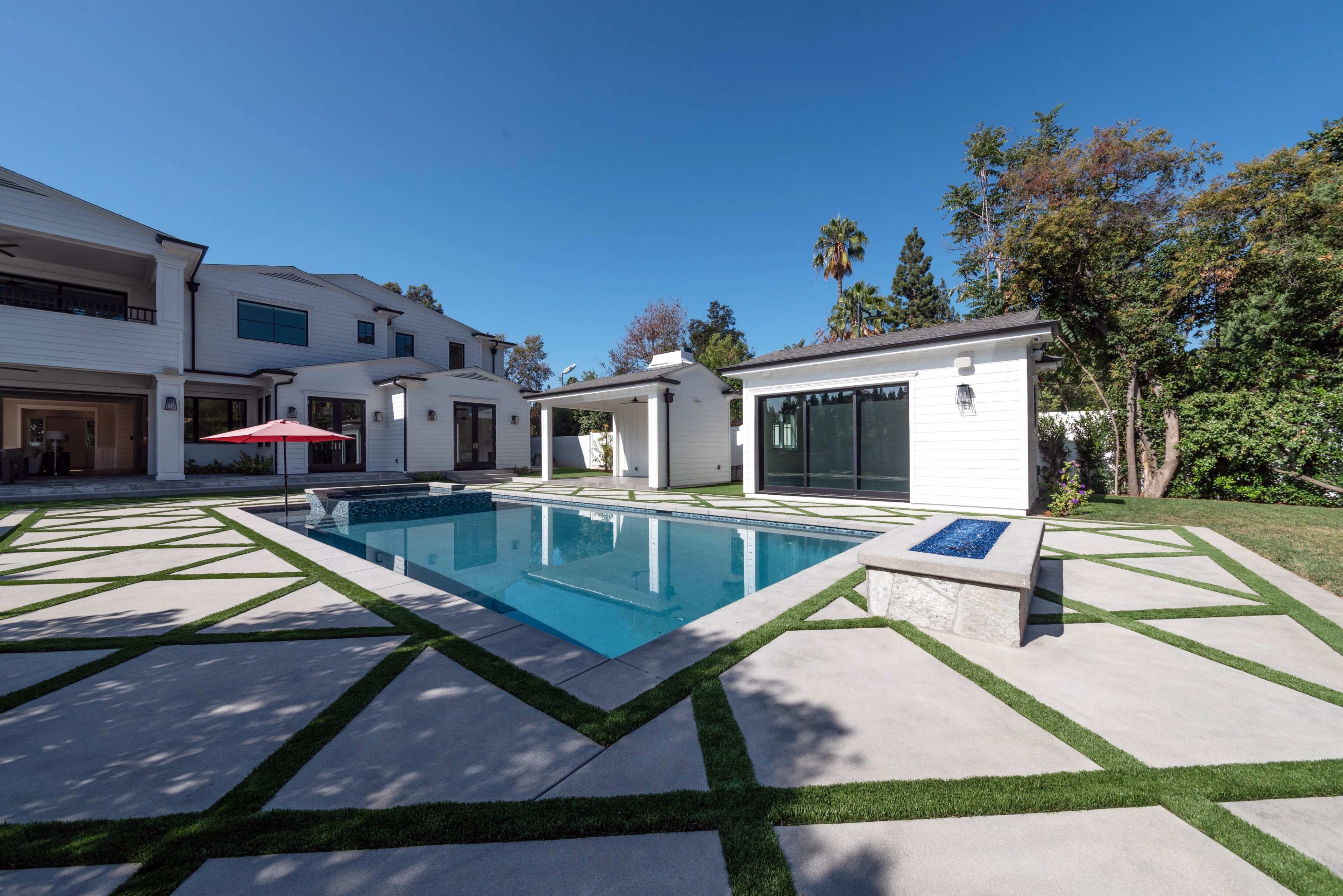 The image shows a modern backyard with a rectangular swimming pool surrounded by concrete and grass pathways, flanked by a pavilion and a covered seating area.