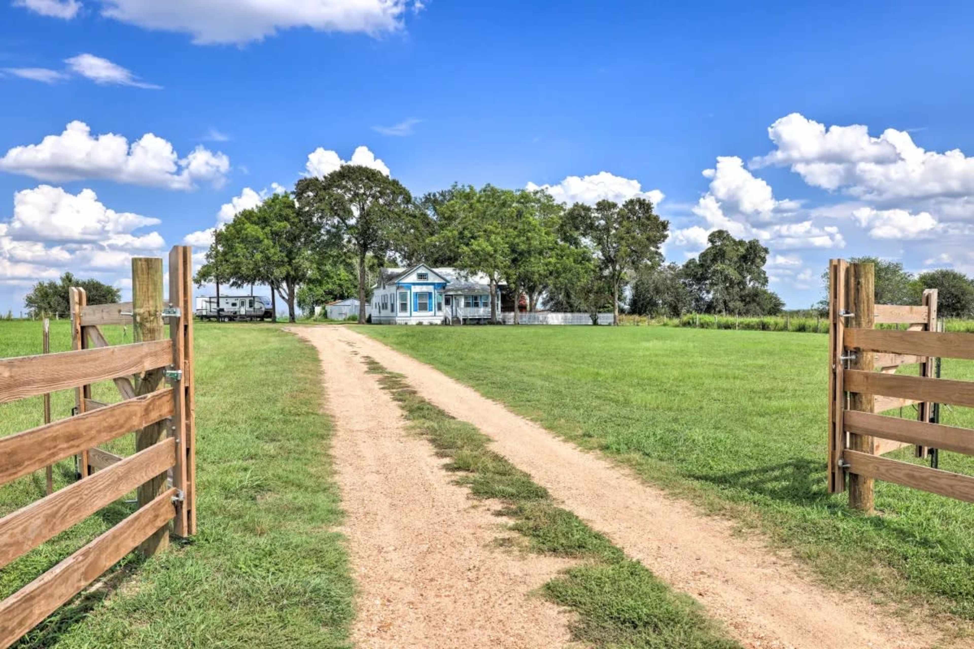A dirt driveway leads from a wooden gate towards a house surrounded by trees and open grassland under a blue sky with clouds.