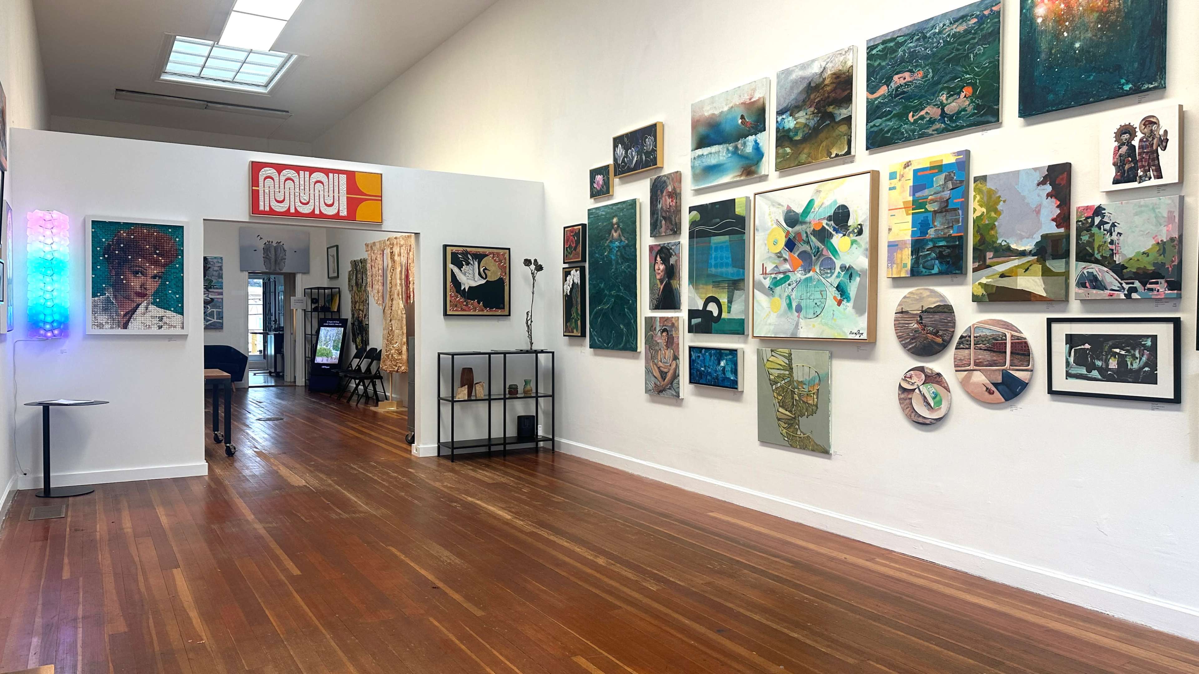 The image shows an art gallery with a long hallway featuring a variety of paintings and artworks displayed on the walls, along with a small table and a person in the background.