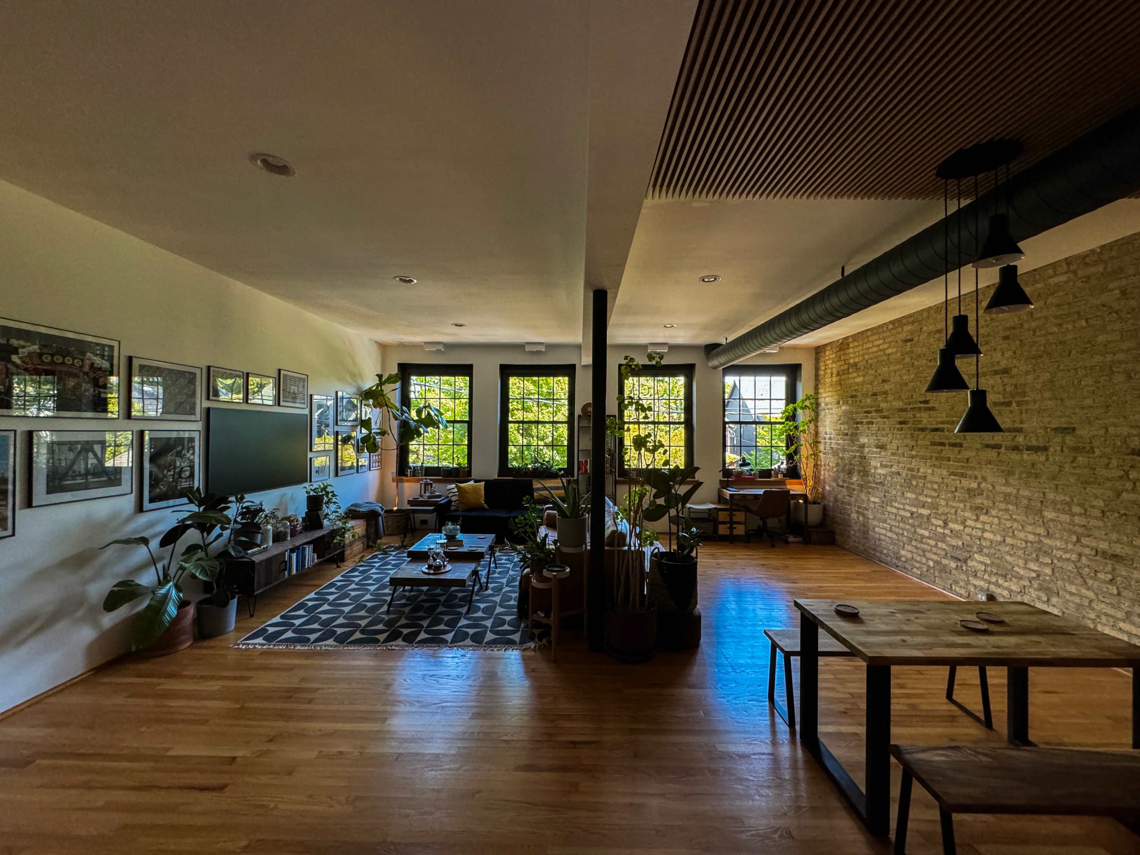 The image shows a spacious living area with wooden flooring, large windows filled with greenery, a patterned rug, and a dining table in the foreground.