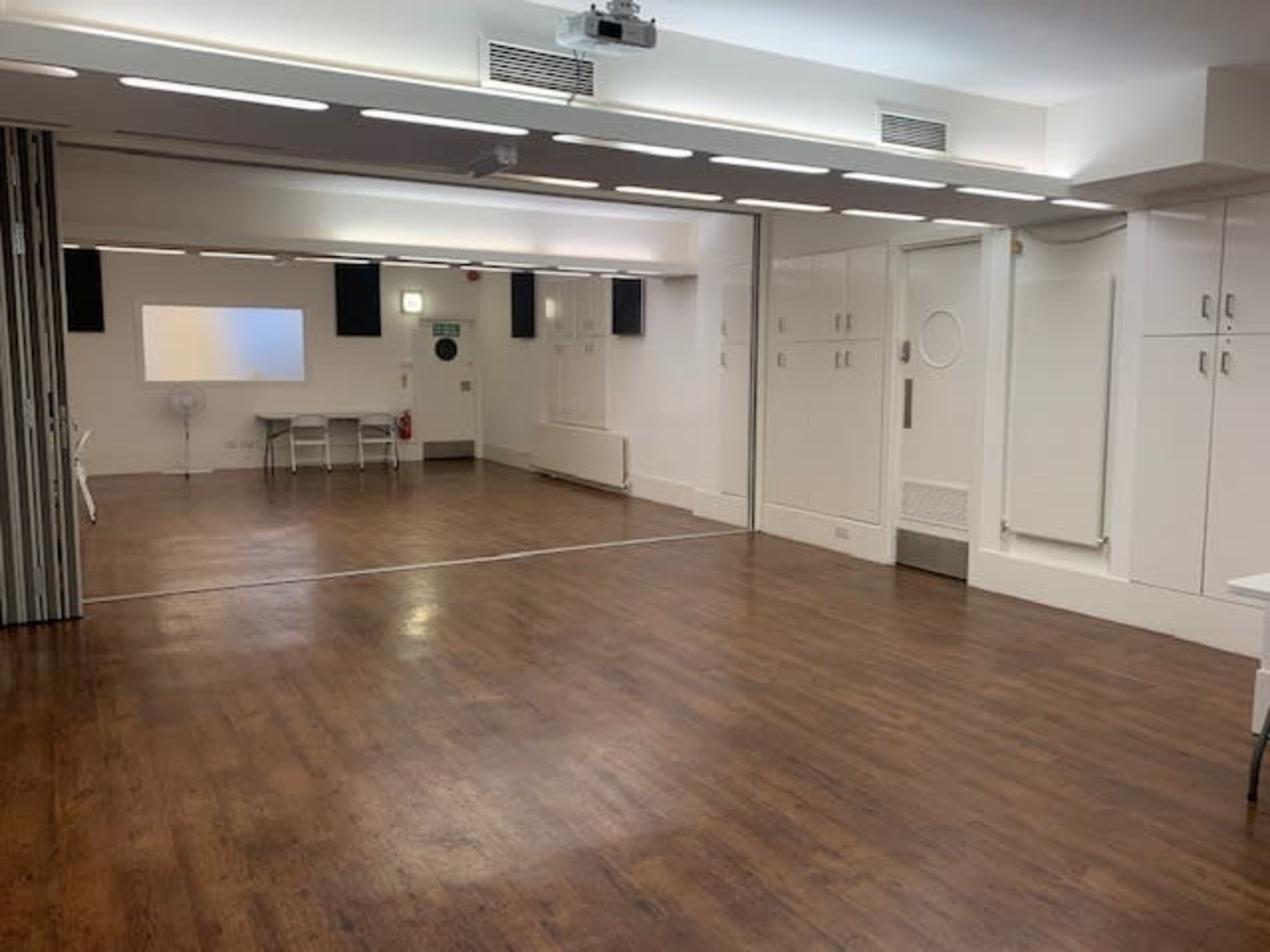 The image shows an empty room with wooden flooring, white walls, a projector screen, and a small table with chairs in the background.