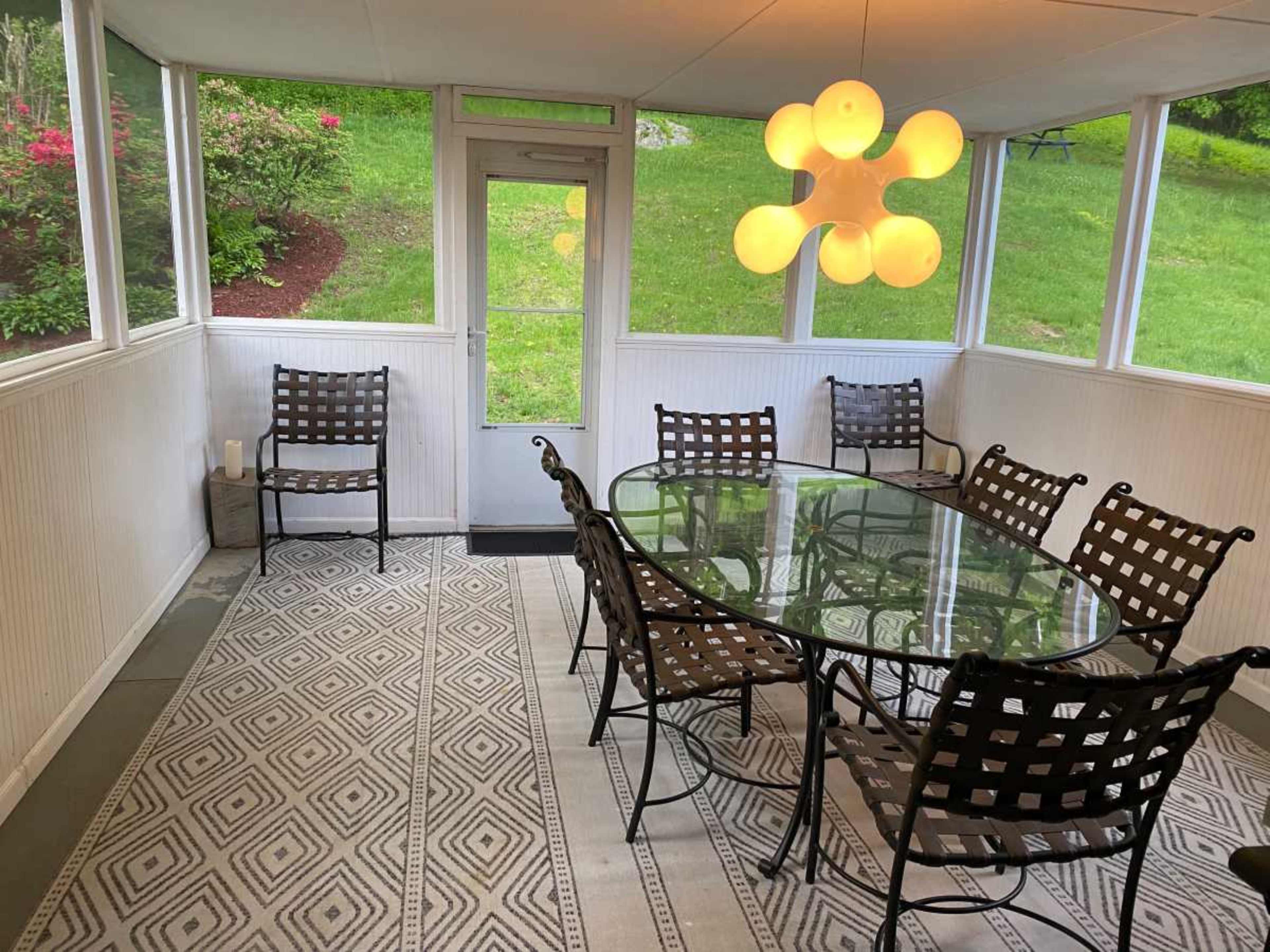 A screened-in porch features a glass dining table surrounded by six metal chairs, with a modern pendant light overhead and greenery visible through the windows.
