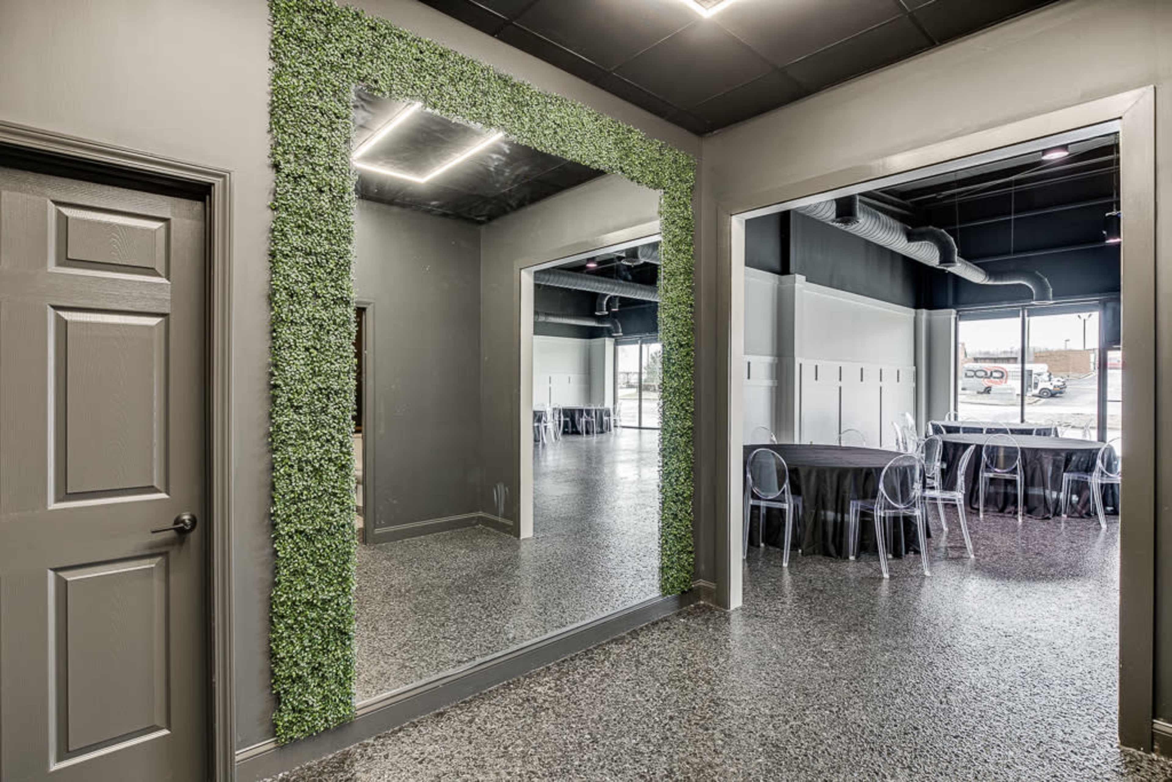 A modern room with a large mirror framed by faux greenery, adjacent to a dining area with black tablecloths and clear chairs.