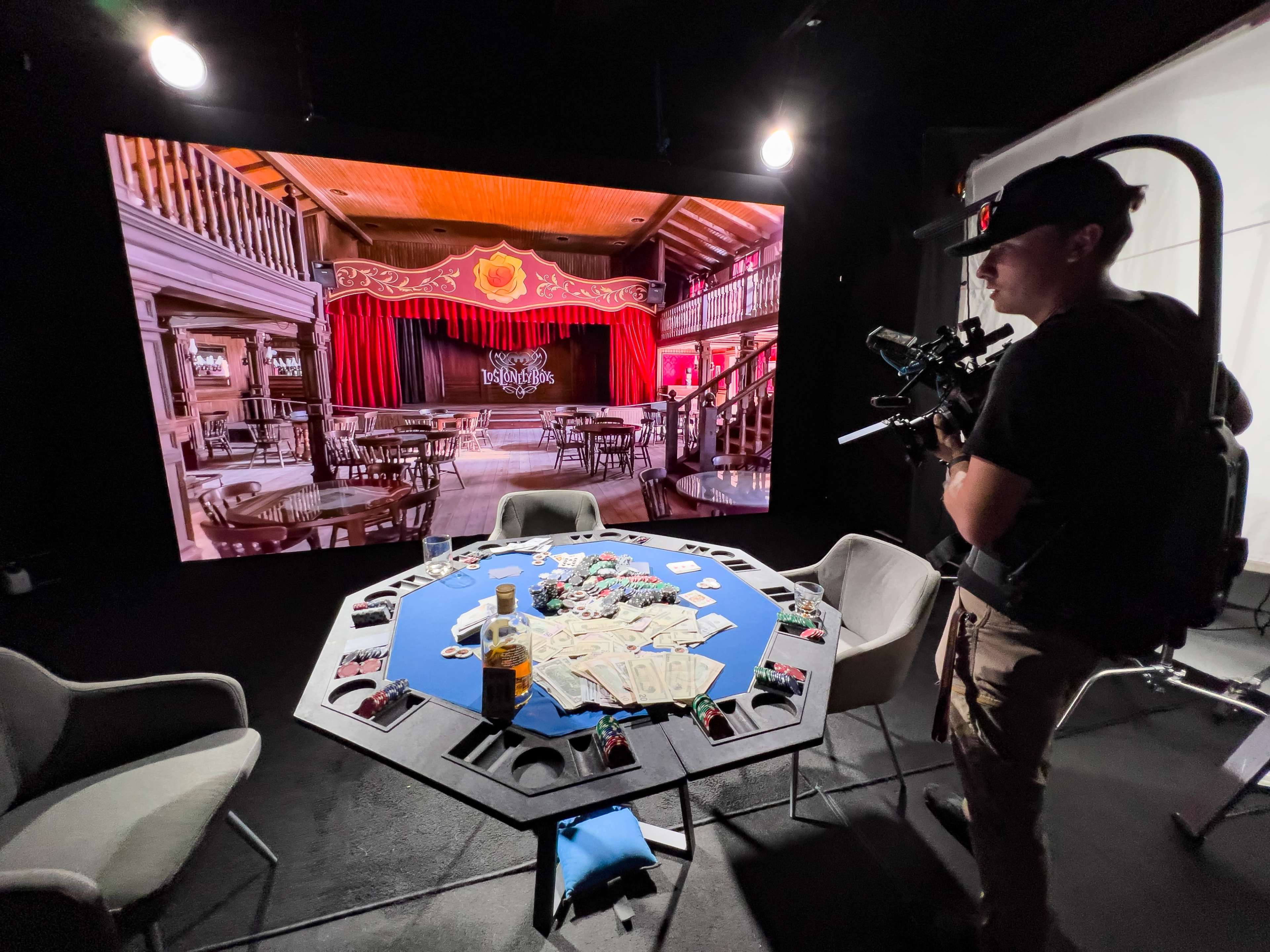 A person stands beside a poker table with money and chips, while a large screen displays a set design of a western-themed bar interior.