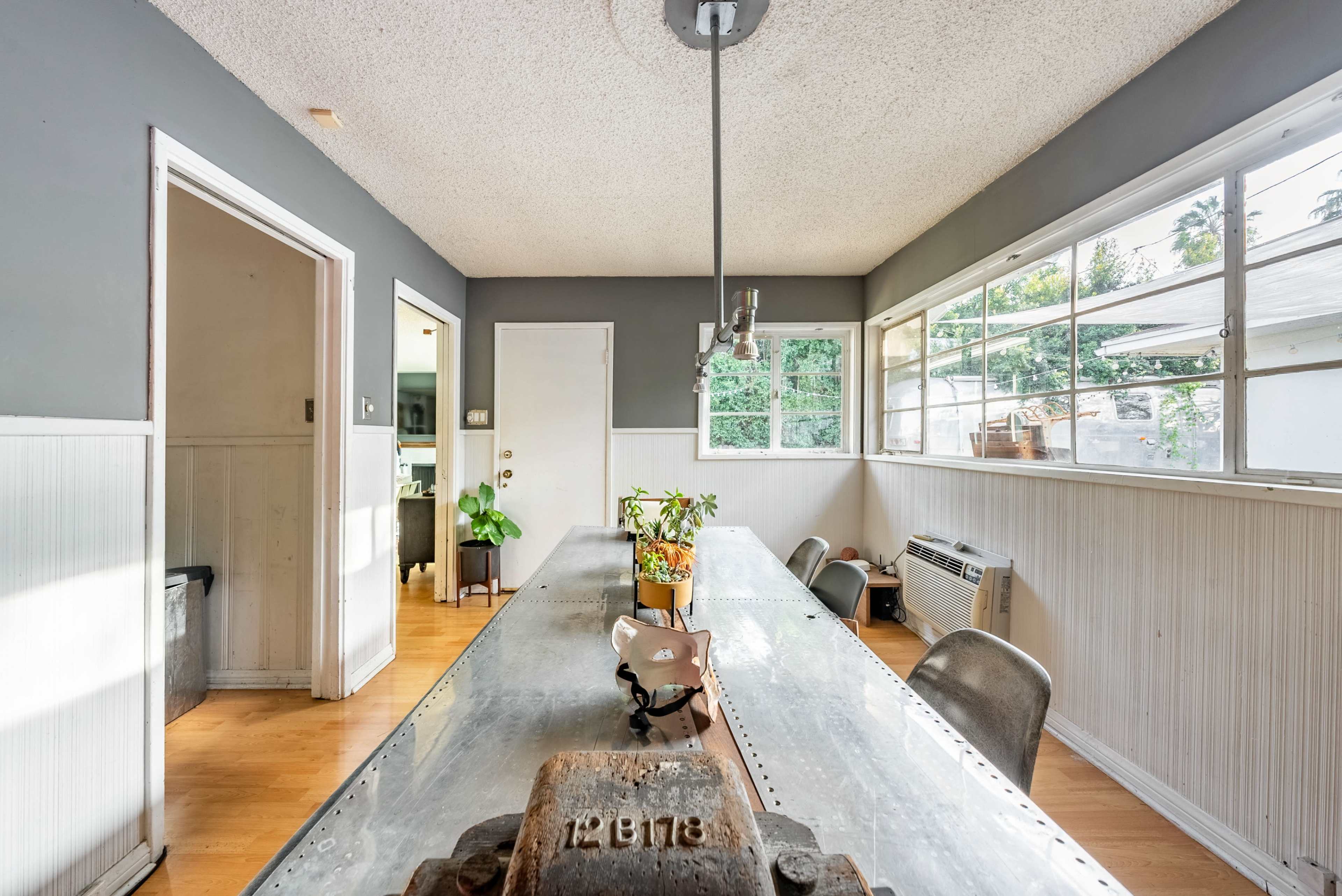 A bright, minimalist dining area with a long metal table, surrounded by steel chairs and windows lining the walls.