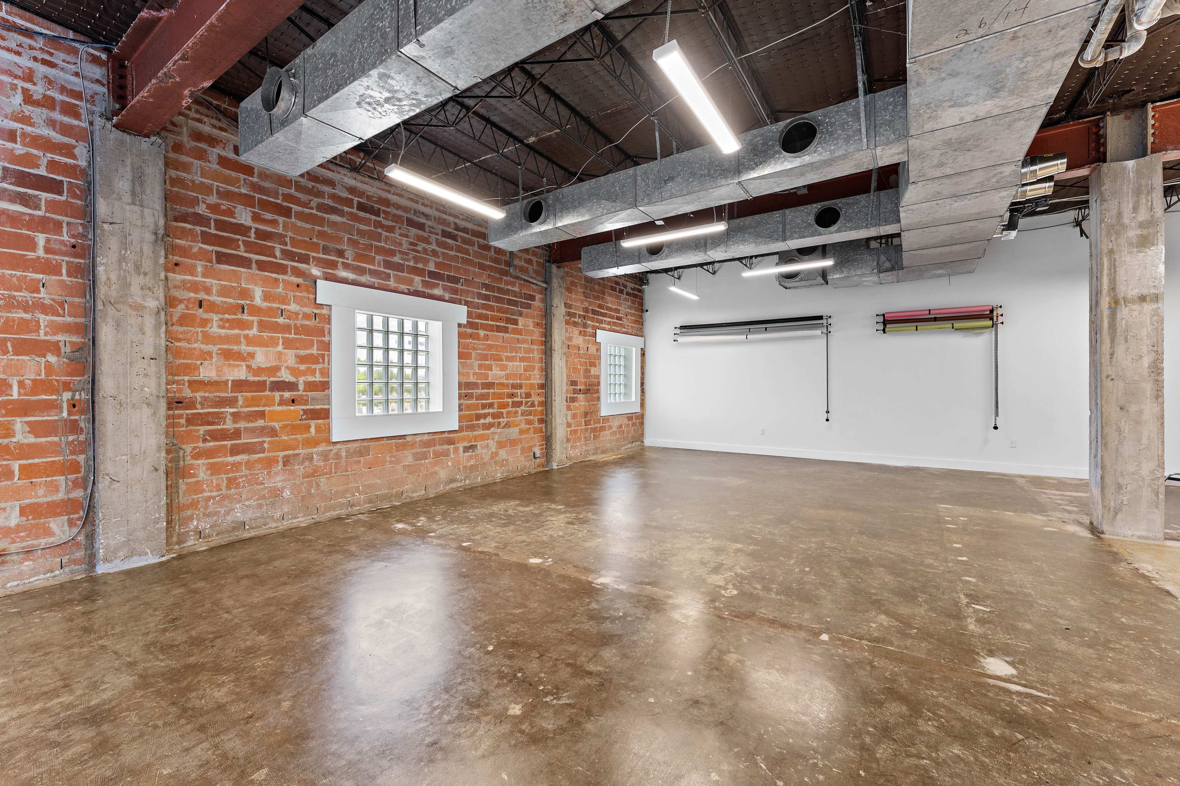 The image shows an empty industrial-style room with exposed brick walls, polished concrete flooring, and overhead ductwork.