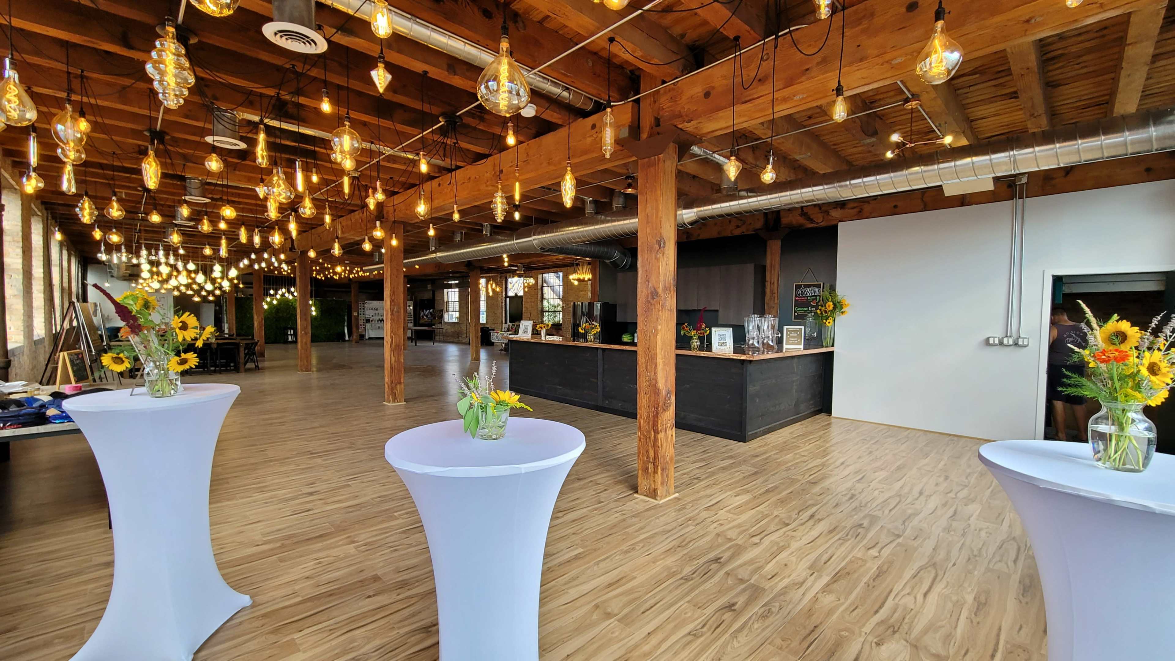 The spacious interior features wooden beams and flooring, with tables adorned with flowers and hanging light bulbs above.