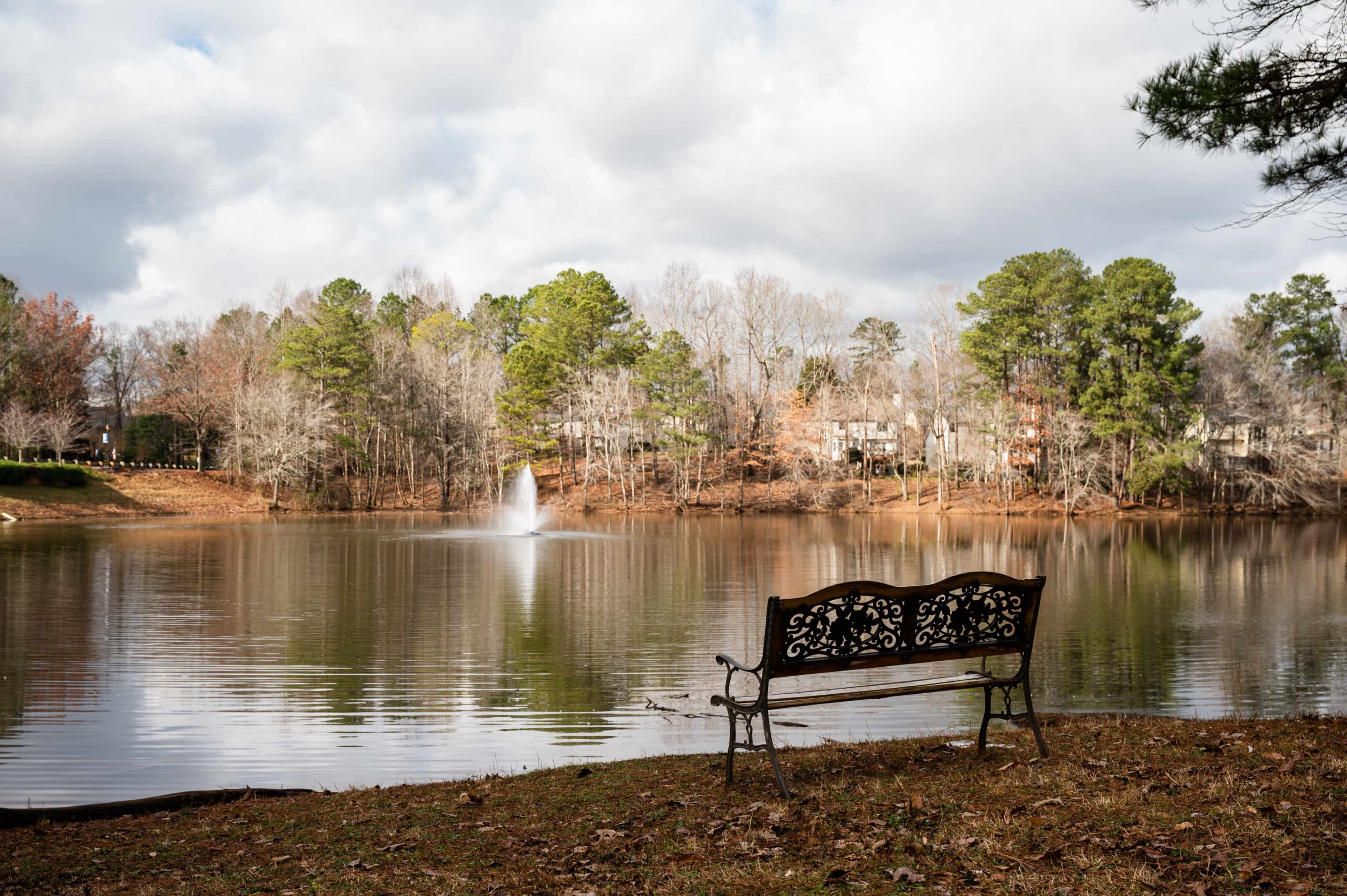 A wrought iron bench sits empty by a calm lake, with a fountain spraying water in the background and trees lining the shore.