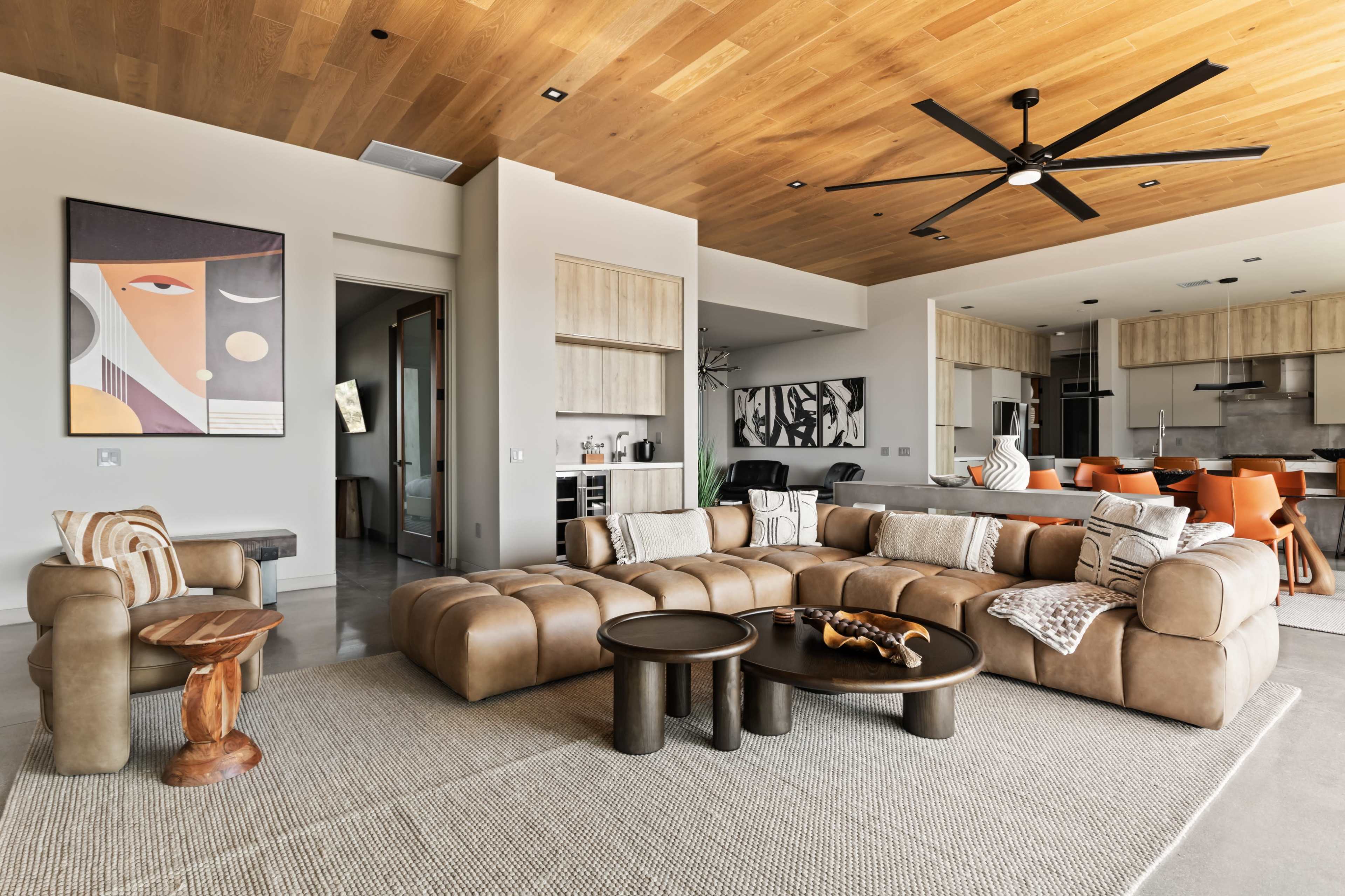 A modern living room features a large brown sectional sofa, wooden ceiling, and a combination of art and furniture styles, creating an inviting space for relaxation.