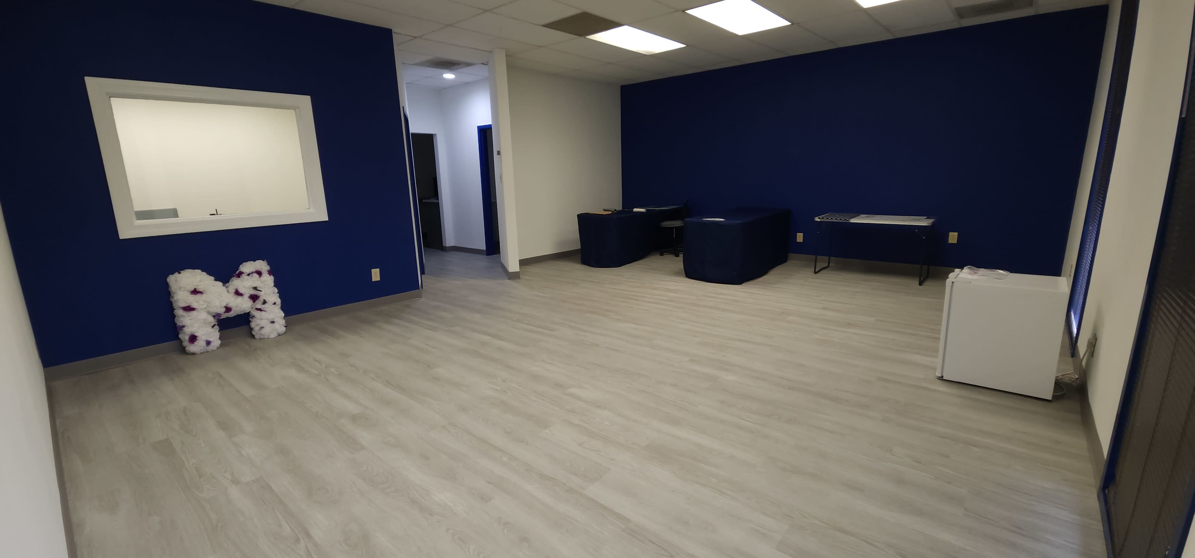 The image shows a sparsely furnished room with a blue wall, light-colored flooring, and a white refrigerator in the corner.