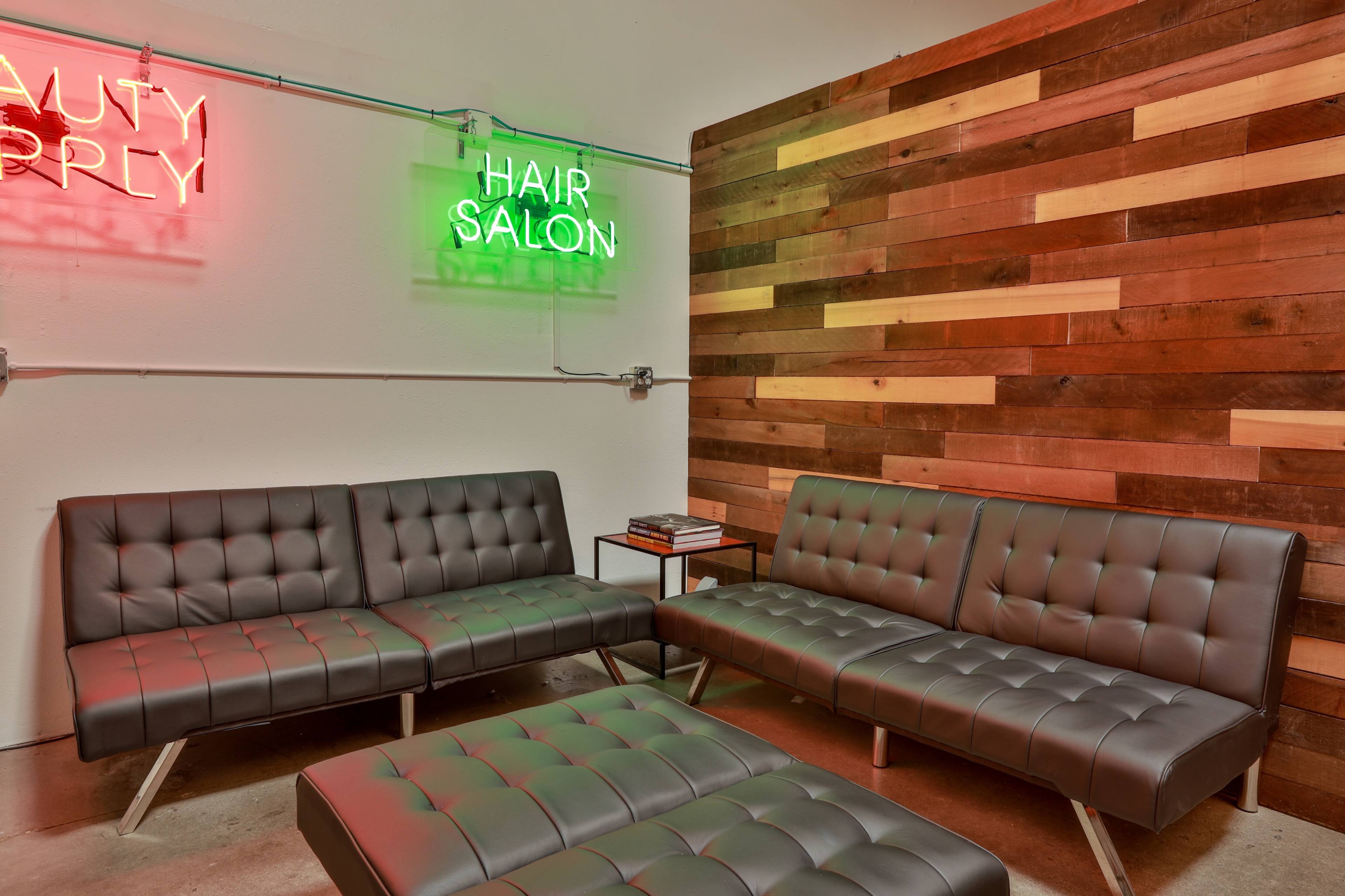 A waiting area furnished with modern gray sofas and a wooden wall, featuring neon signs that read "BEAUTY SUPPLY" and "HAIR SALON."