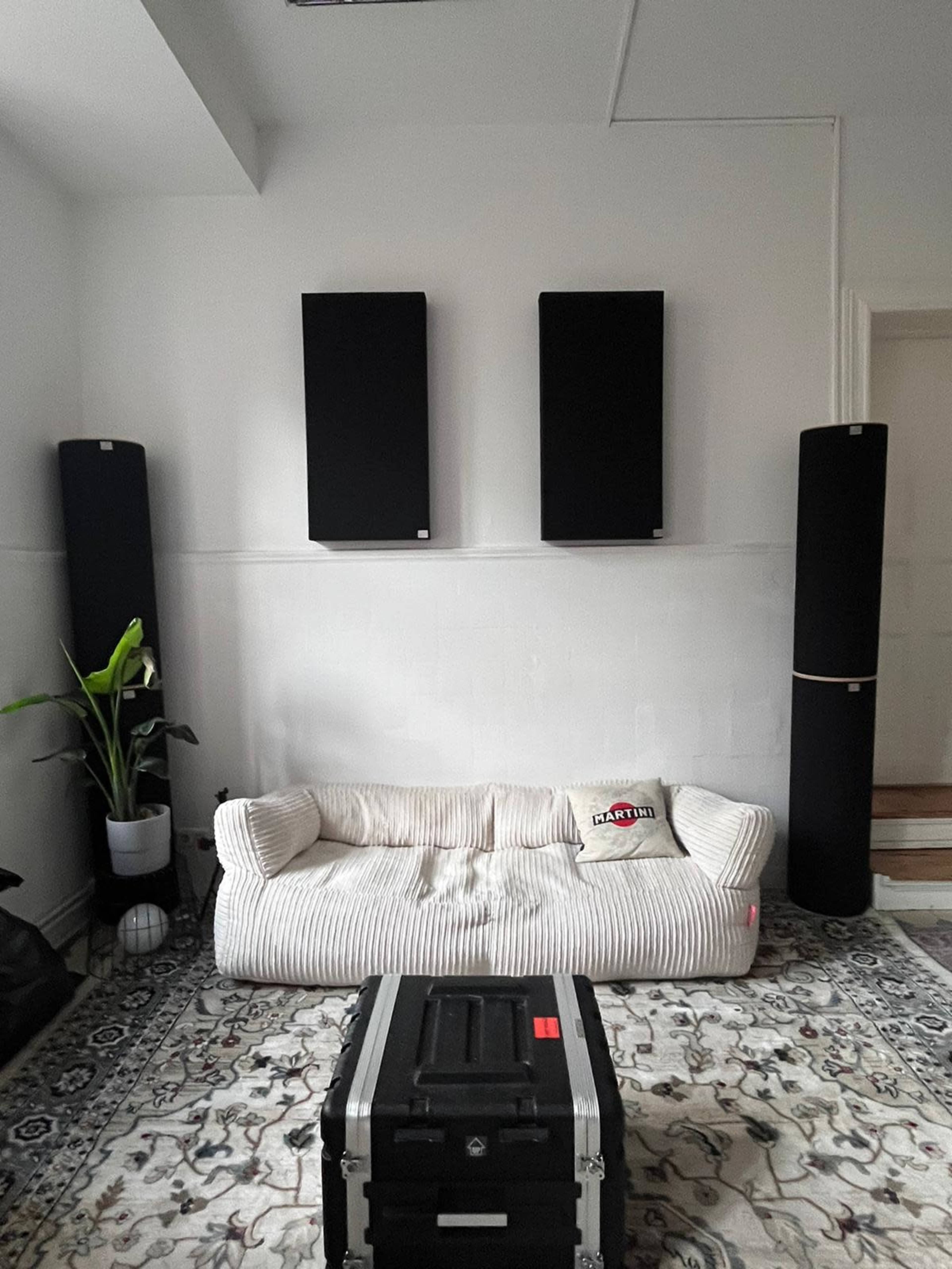The image shows a minimalist room with a large, striped couch, two tall speaker-like installations on the walls, a potted plant, and a black storage case on a patterned rug.