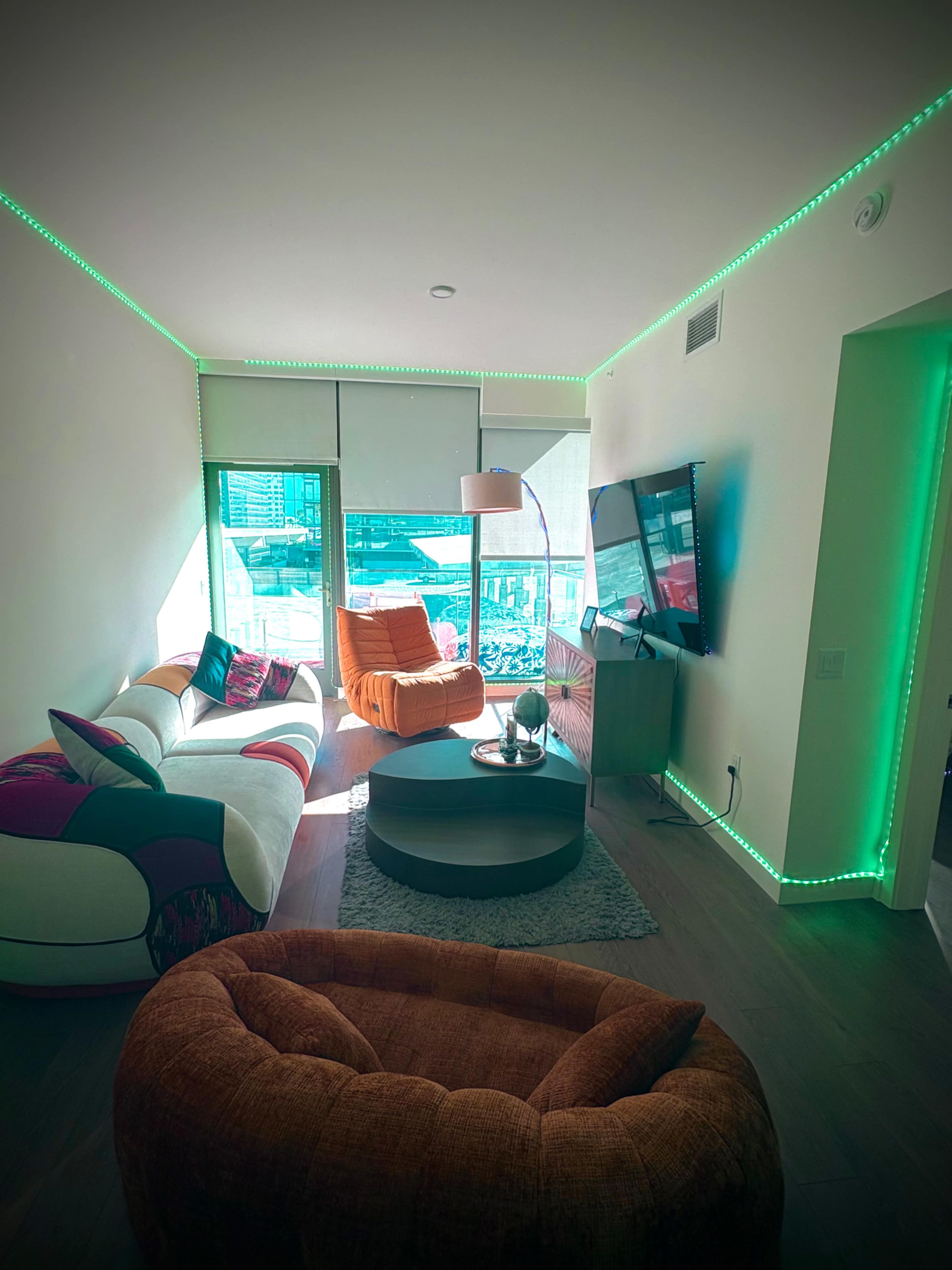 The living room features a sectional sofa, two orange armchairs, a round coffee table, and a mounted television, all illuminated by green LED lights along the walls.