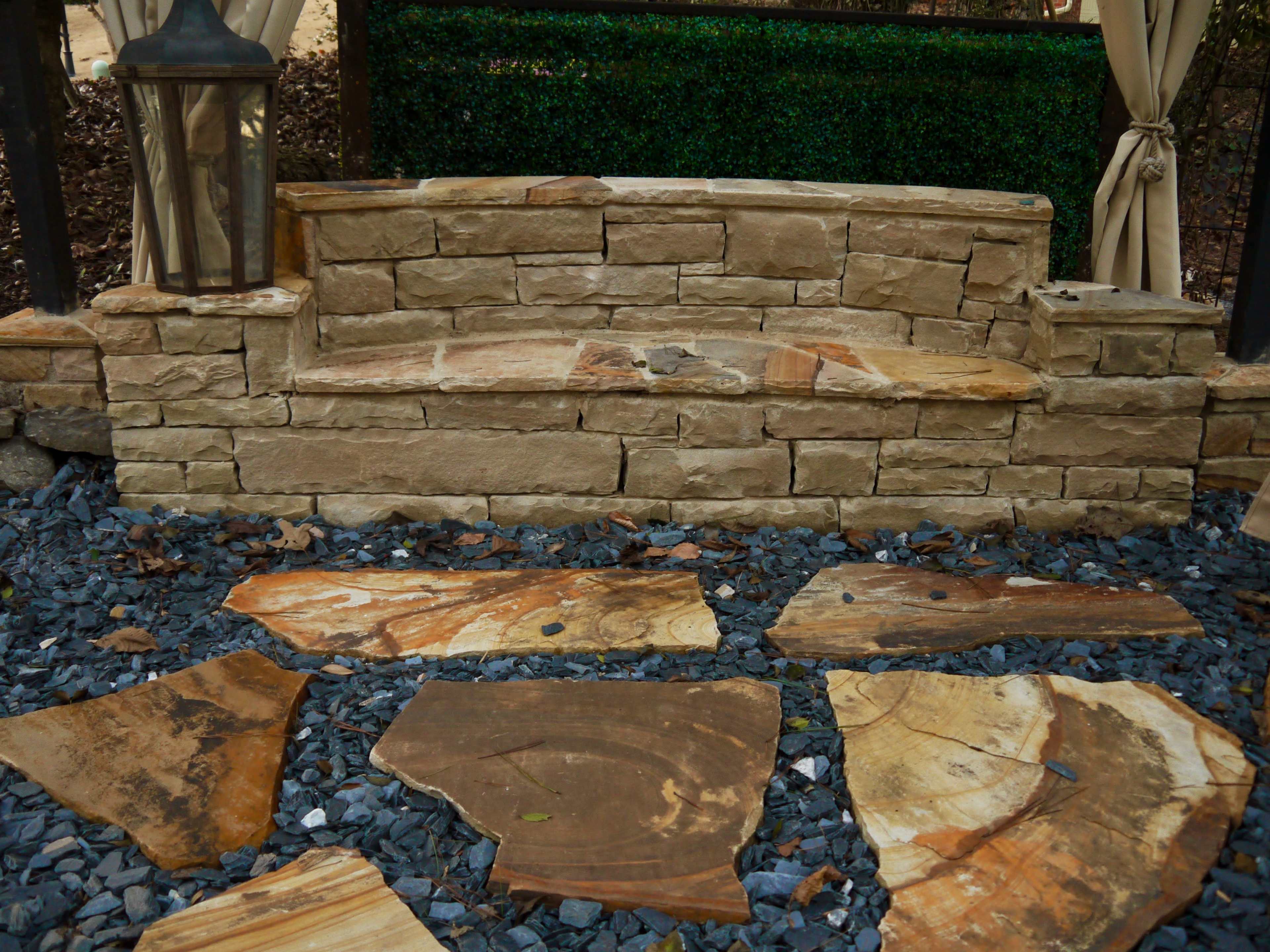 A stone bench made of stacked stone blocks, positioned on a bed of dark gravel and surrounded by irregularly shaped stone slabs.