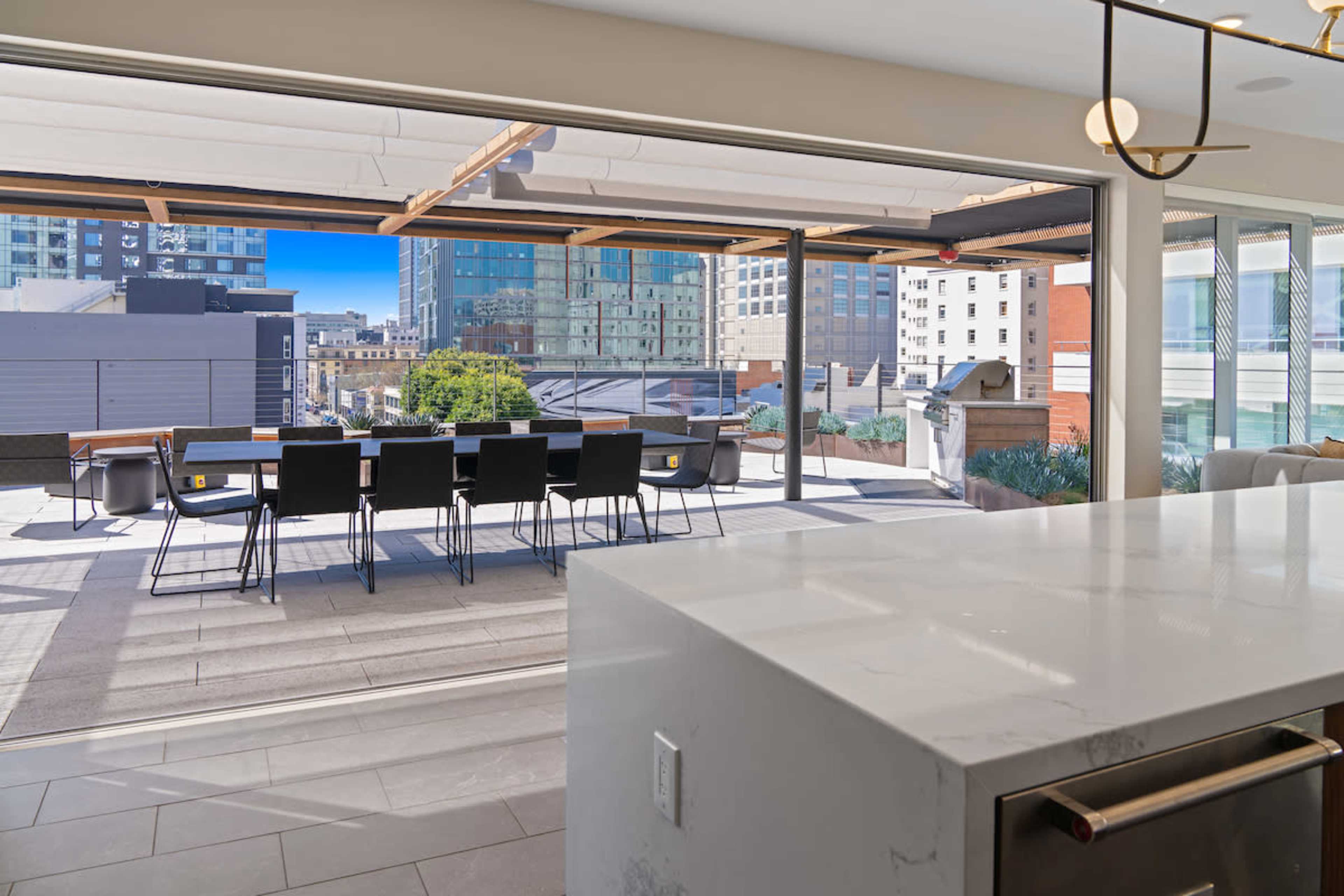 The image shows a modern indoor space with a large open window revealing a patio featuring a dining table and seating against a city skyline backdrop.