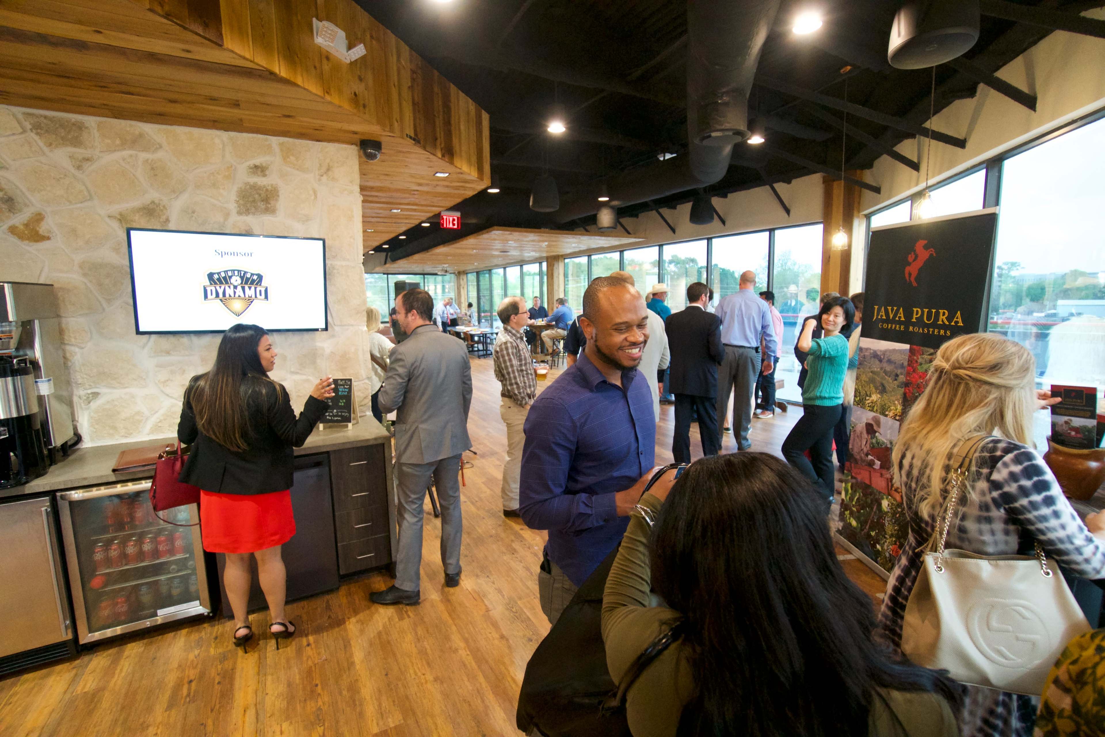 A group of people mingling in a modern event space, featuring a refreshment area and large windows with views of the outside.
